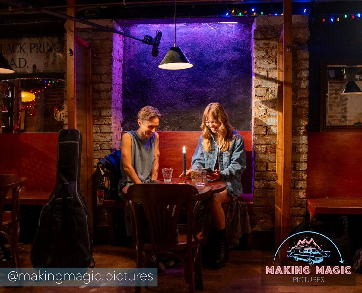 Magic and Jo are sitting at a small wooden table in a cozy, dimly lit bar. Jo is holding a phone, and there's a lit candle between them. They are smiling and appear to be enjoying a conversation. The background features exposed brick walls, colorful 