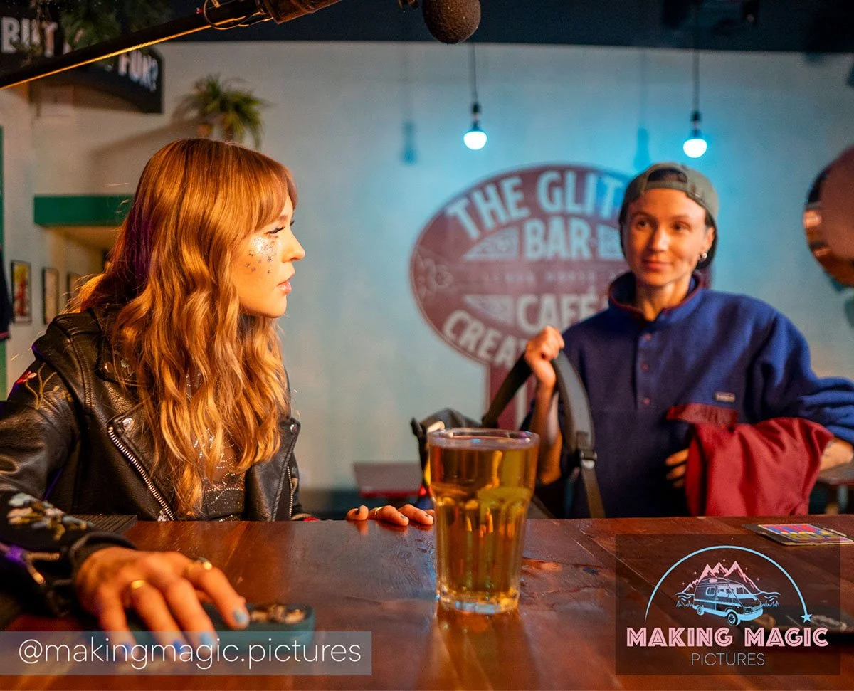 Two people sit at a bar table in a lively cafe, one with long wavy hair and the other with short hair and a baseball cap, engaged in conversation with a glass of beer on the table. The cafe has a neon sign and decorative lighting.