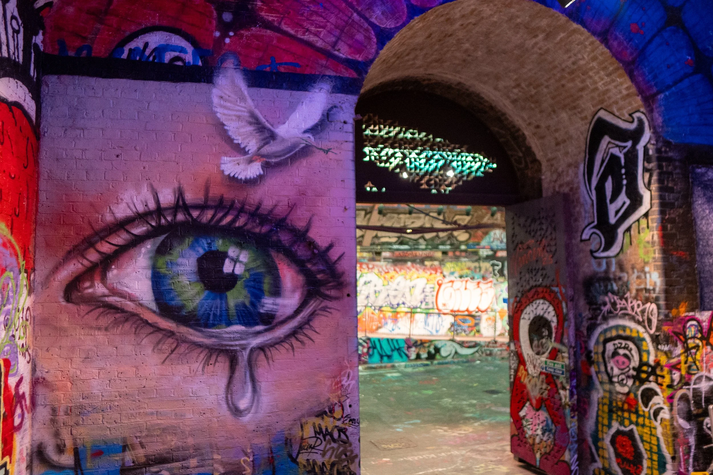 Graffiti art of a large eye with a tear on a brick wall, a flying bird above, and vibrant, colorful spray-painted murals inside an arched brick passageway.