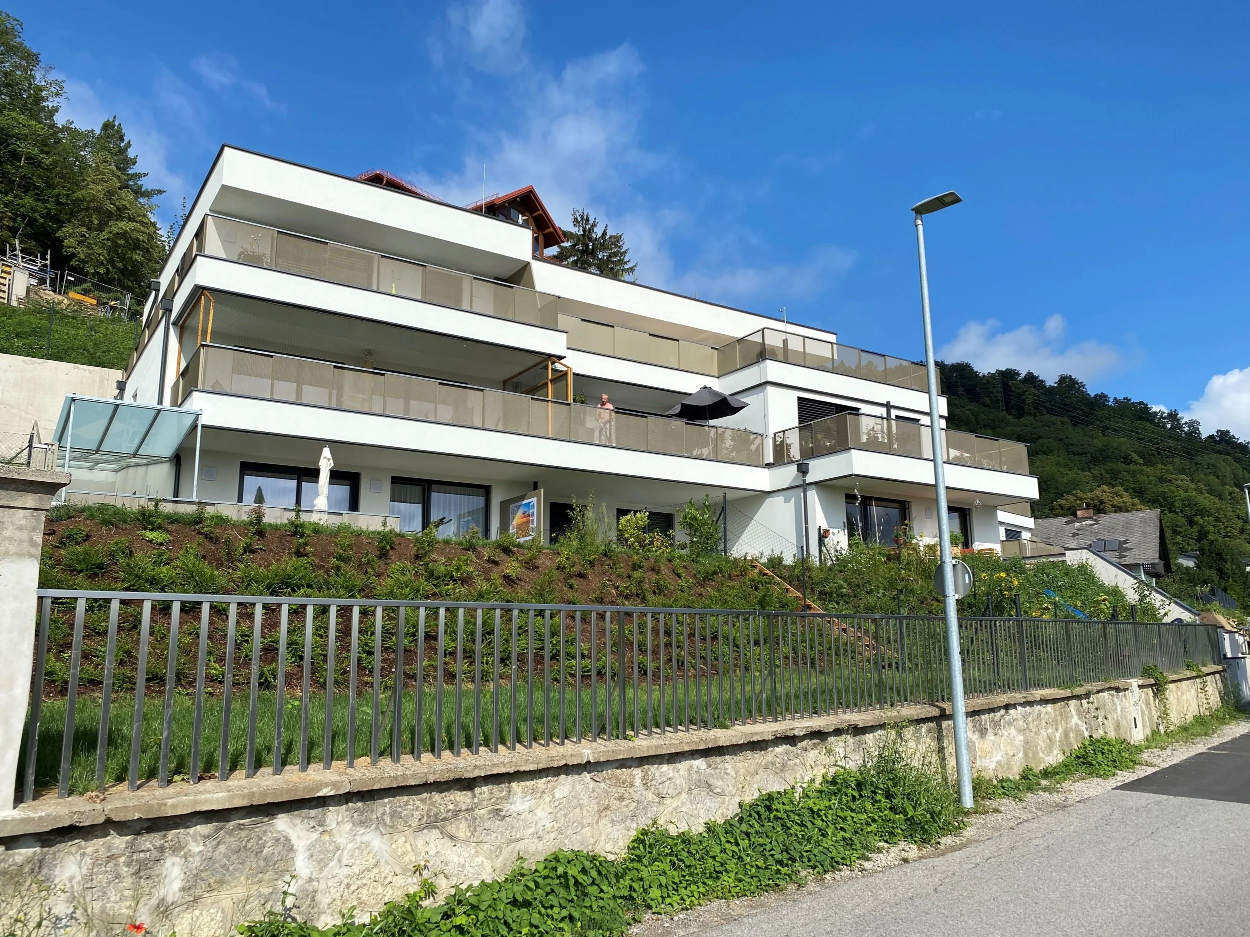 Modern white multi-story house with multiple balconies, large glass doors, and a sloped roof, situated on a hillside with greenery and trees in the background.