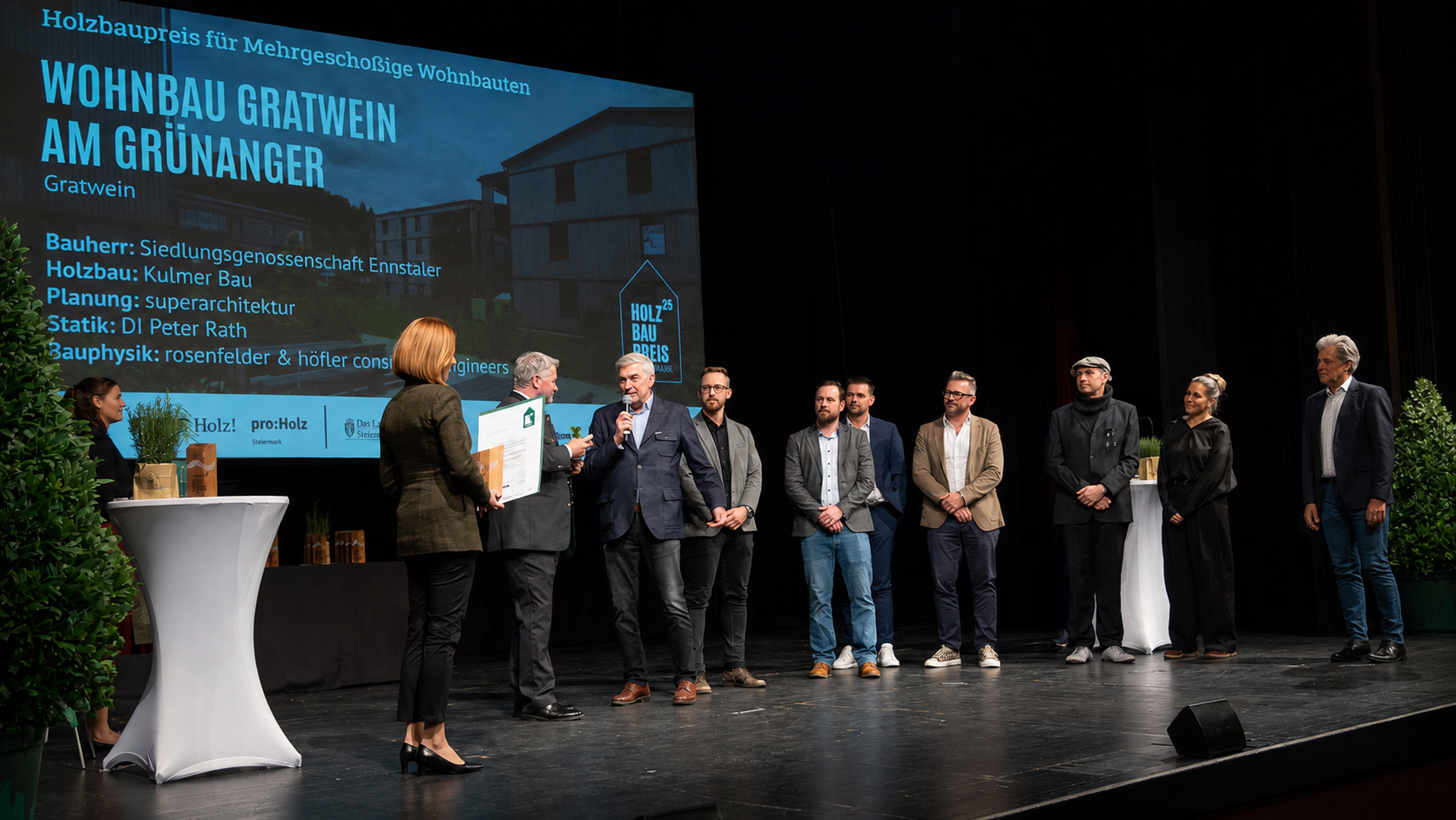 Group of people on stage during an award ceremony, with a large screen in the background displaying information about a housing project in Graz. The scene includes a woman presenting an award to a man, and several men standing in line, some holding papers or awards. The stage is decorated with plants and high tables.