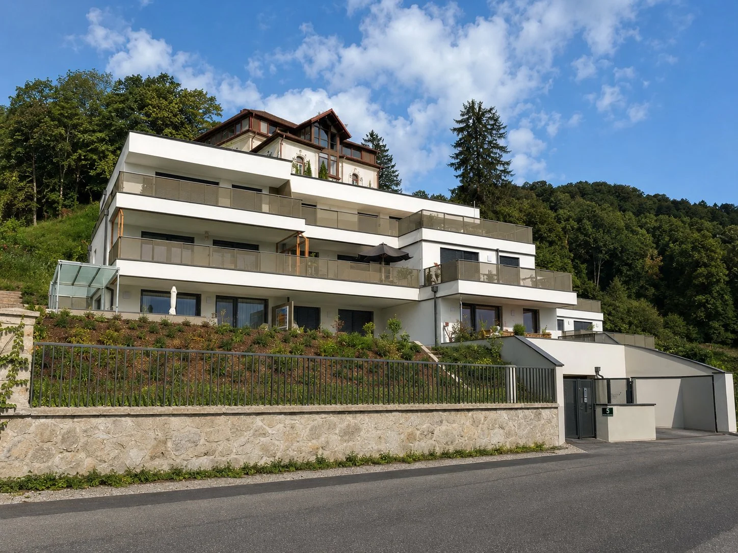 Modern multi-level white building with balconies and glass railings, situated on a hillside with a forested area and blue sky in the background.