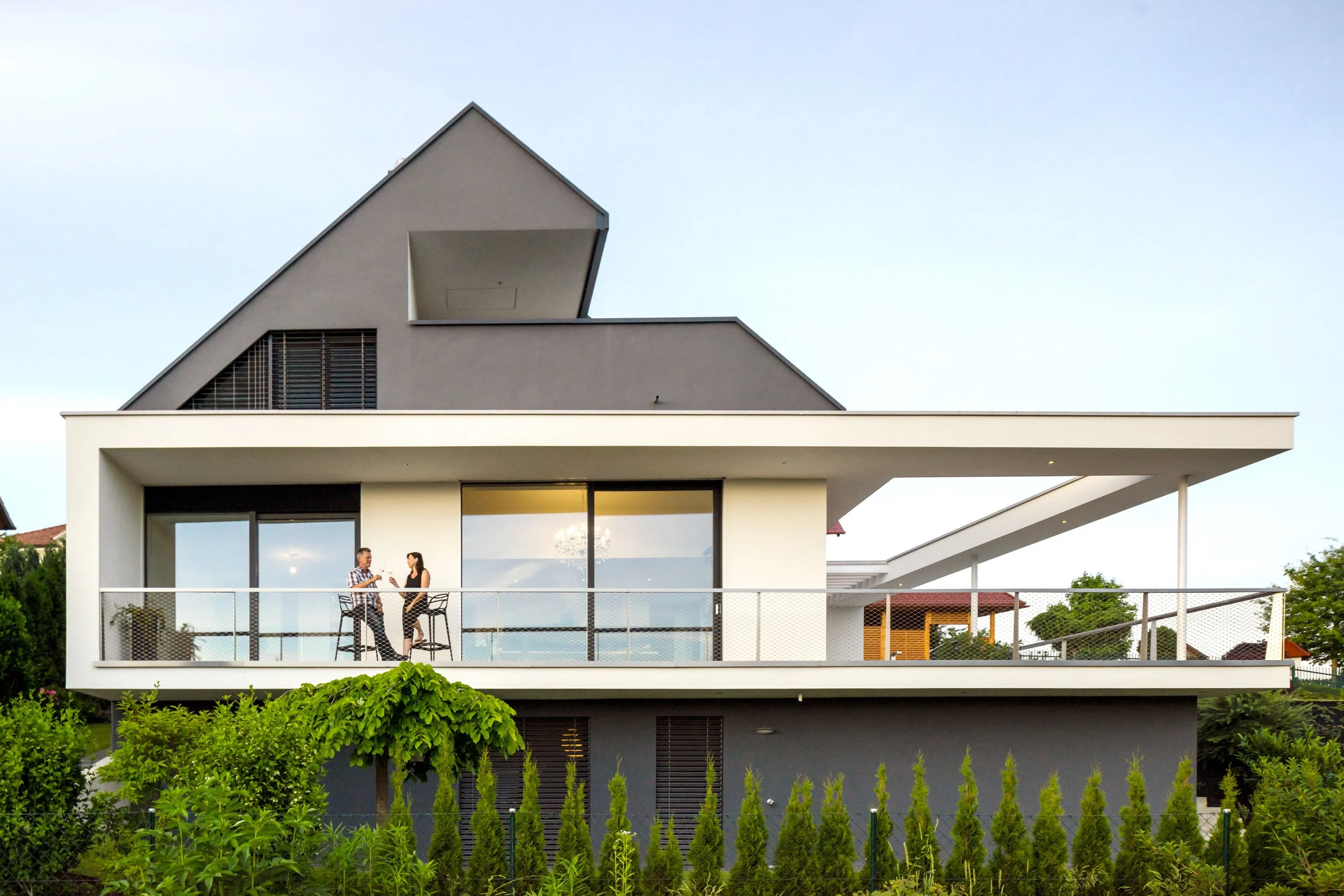 Modern house with a large balcony, two people standing and talking, lush green plants in the foreground, and a clear blue sky.