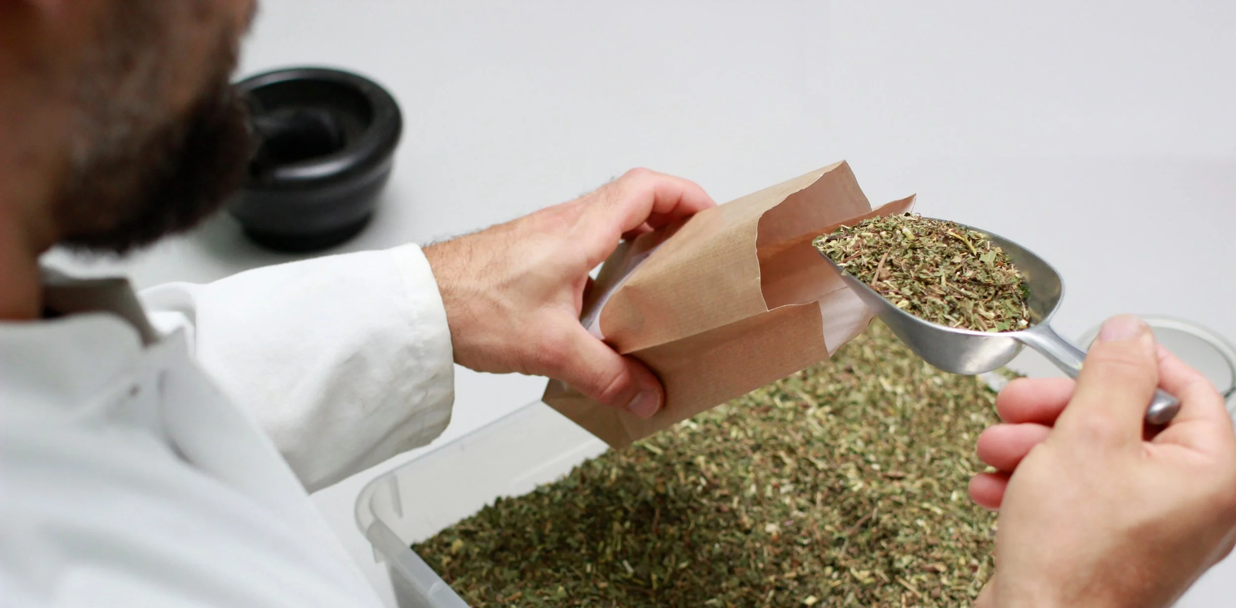 Mann schneidet getrocknetes Kräutermischung mit einer Schaufel aus einer transparenten Box, die auf einem weißen Tisch steht.