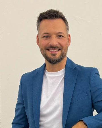 Ein Mann mit Barton-Schläfen, dunklem Haar und Bart, trägt einen blauen Anzug und ein weißes T-Shirt, steht vor einer weißen Wand und lächelt.