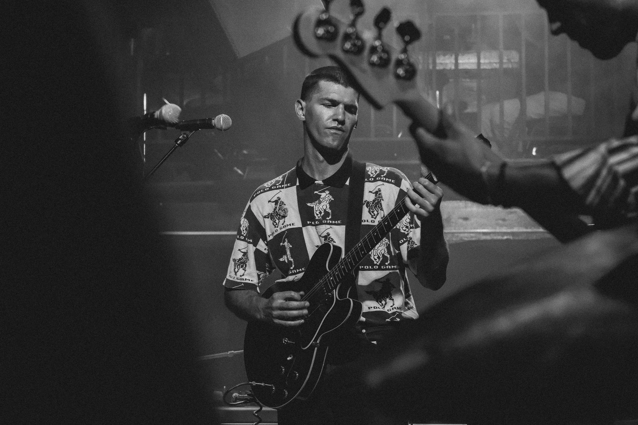 A musician playing an electric guitar on stage, wearing a patterned shirt with a logo, with a microphone and another person holding or adjusting the guitar, in black and white.