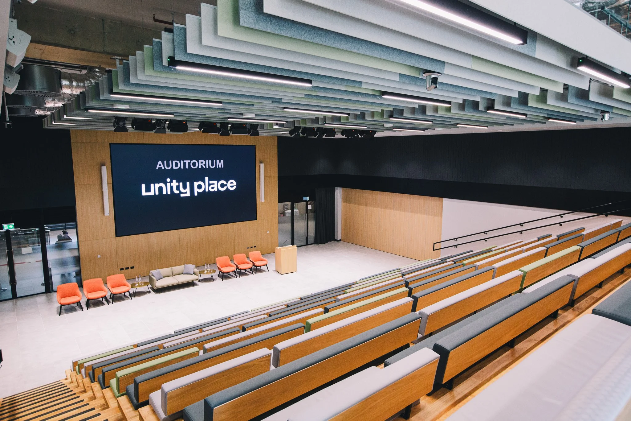 An auditorium with tiered seating in various colors, a stage area with orange chairs and a beige sofa, a large digital screen displaying 'Auditorium Unity Place,' and a ceiling with modern acoustic panels and suspended lights.