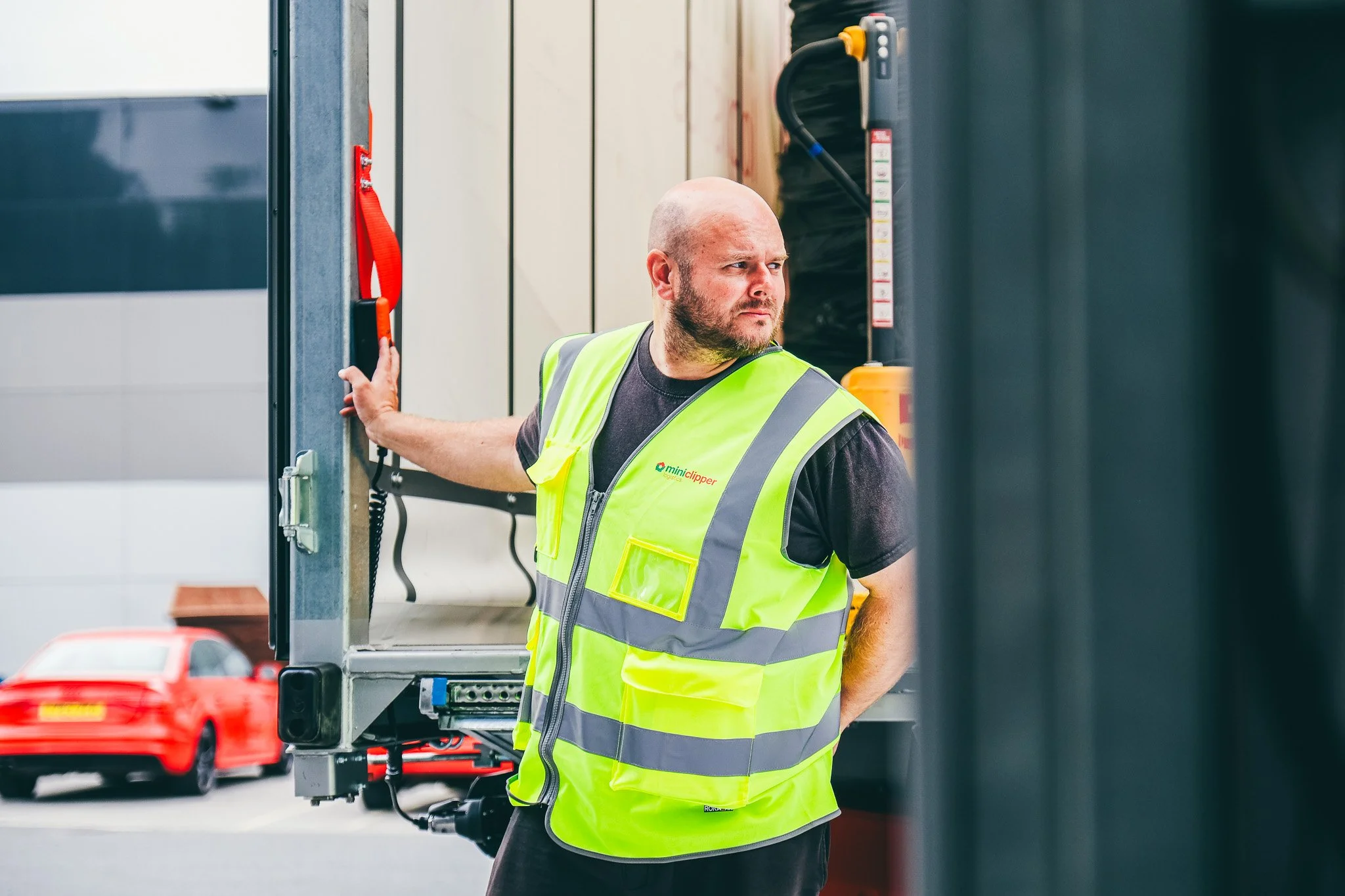 A man with a bald head and beard wearing a high-visibility vest operating a vehicle lift or hoist in a parking lot.