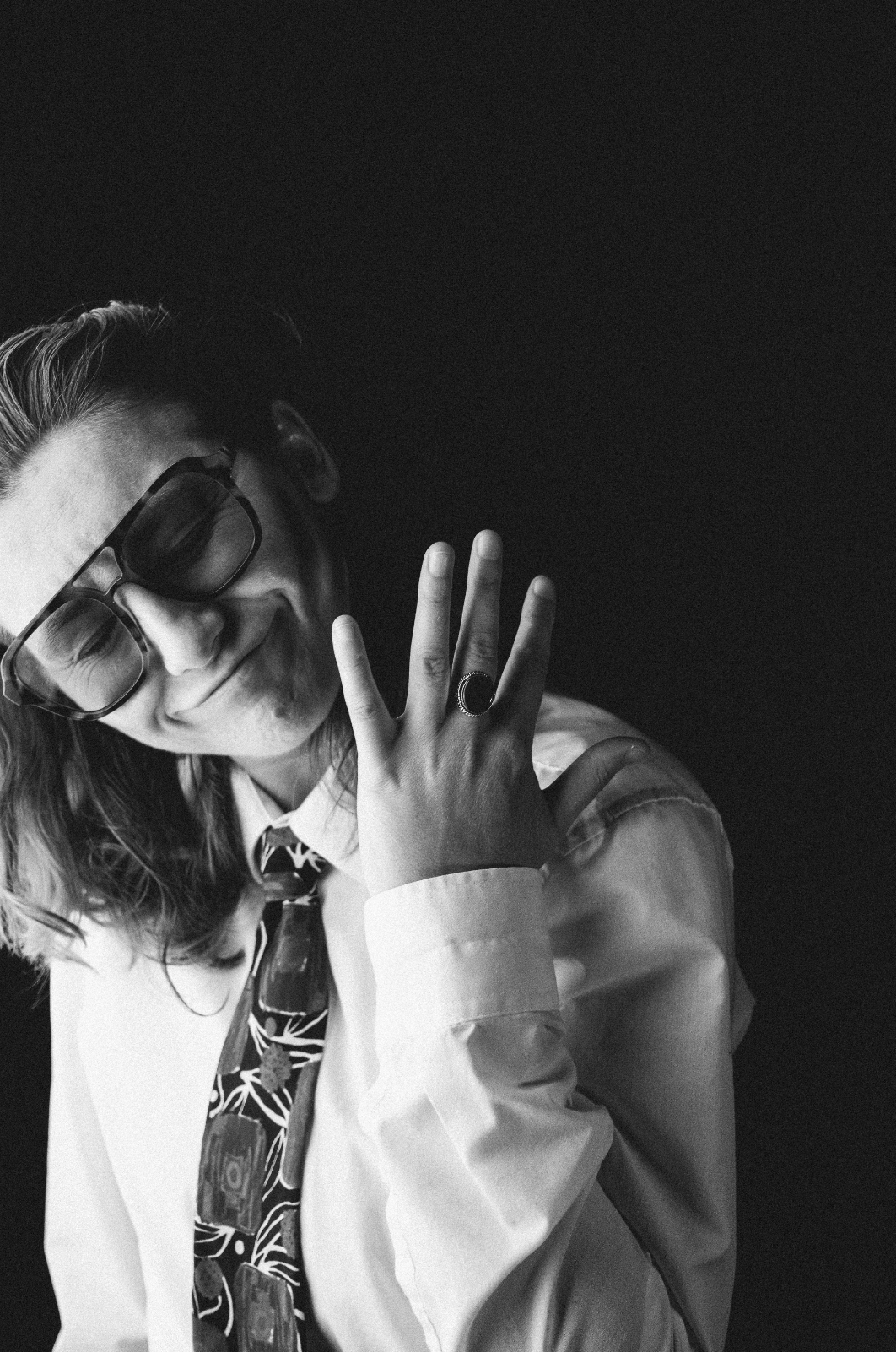 Black and white portrait of a person wearing glasses, a patterned tie, a white shirt, and a ring, smiling and posing with one hand near their face against a dark background.