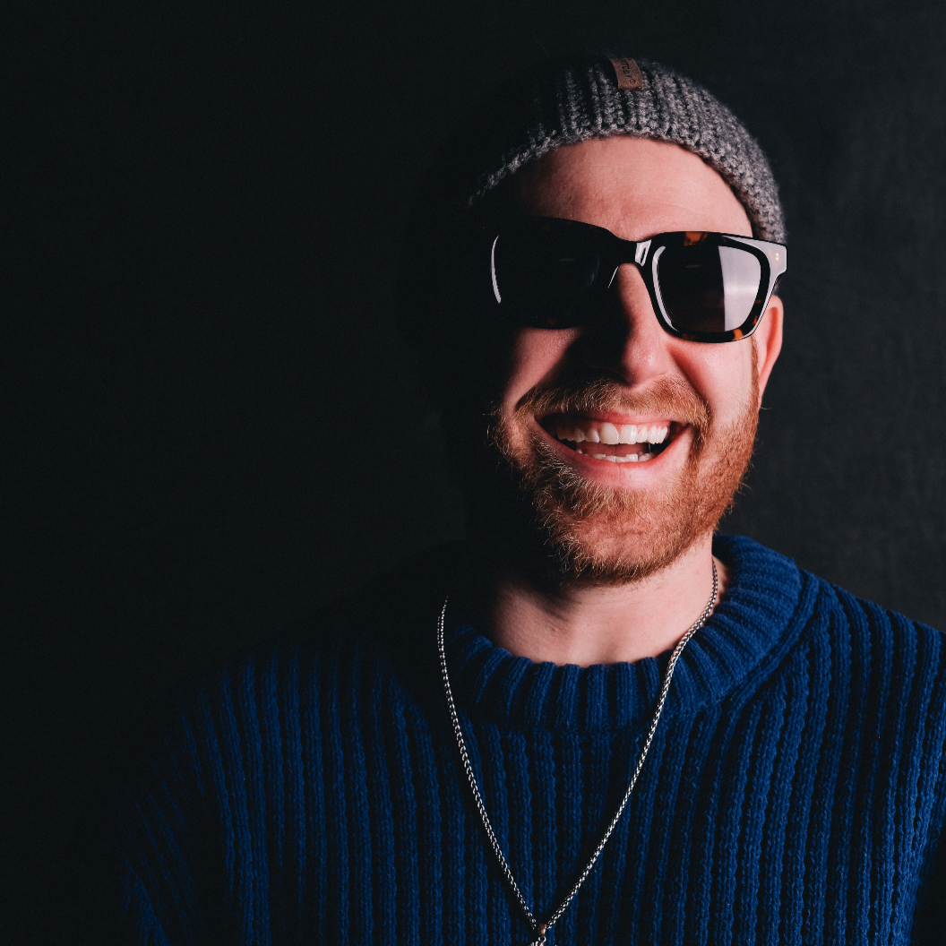 A man with a beard wearing sunglasses, a gray knit beanie, a blue sweater, and a silver chain, smiling against a black background.