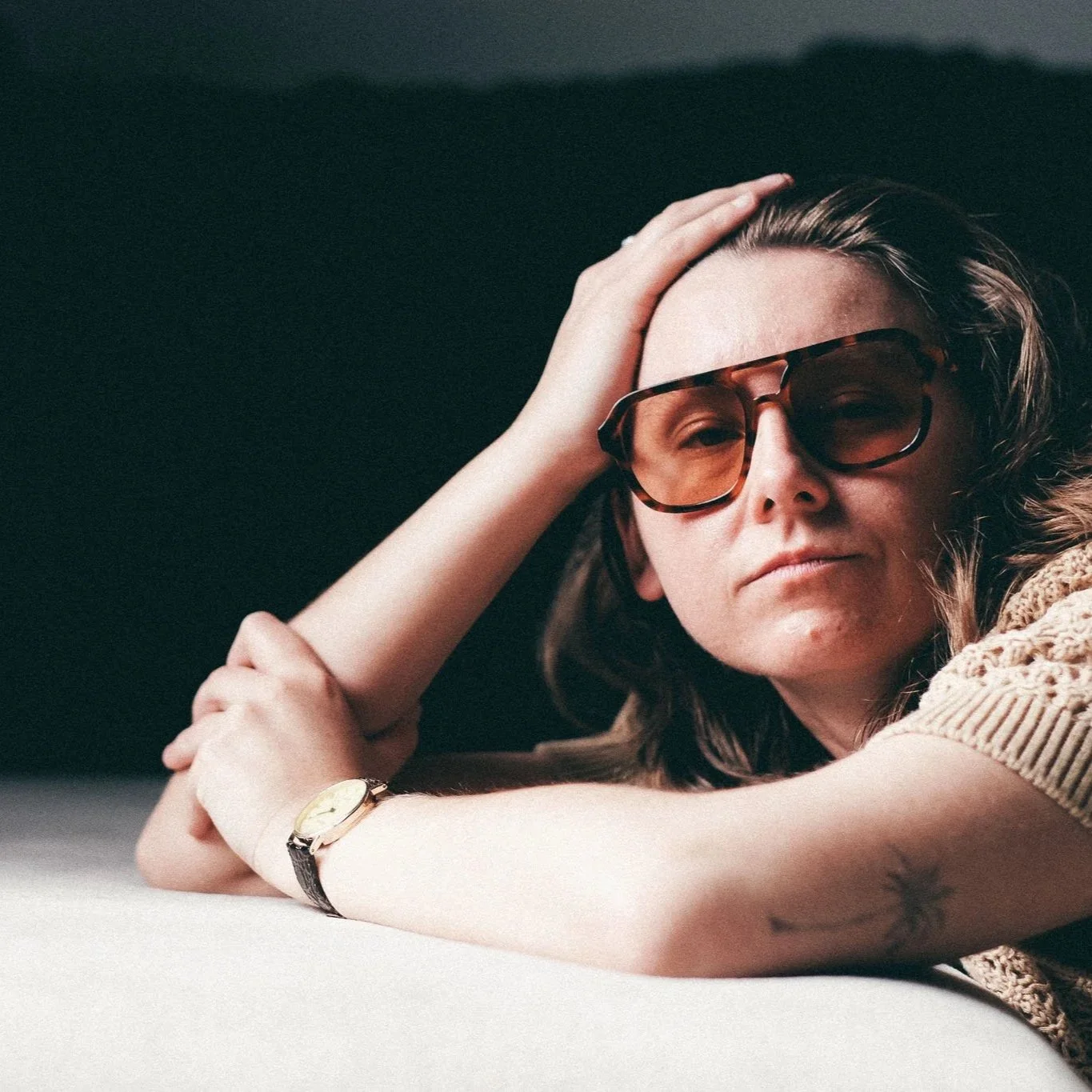 A woman with long brown hair wearing tortoise shell sunglasses and a beige knitted sweater, resting her head on her hand and looking at the camera, with a black background.