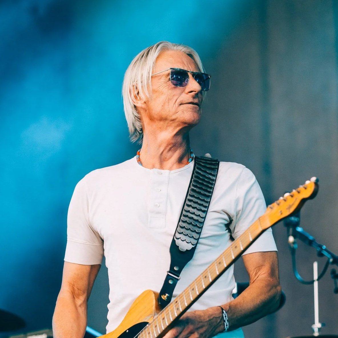 An older man with white hair wearing sunglasses and a white shirt, holding an electric guitar, standing against a blurred blue background.