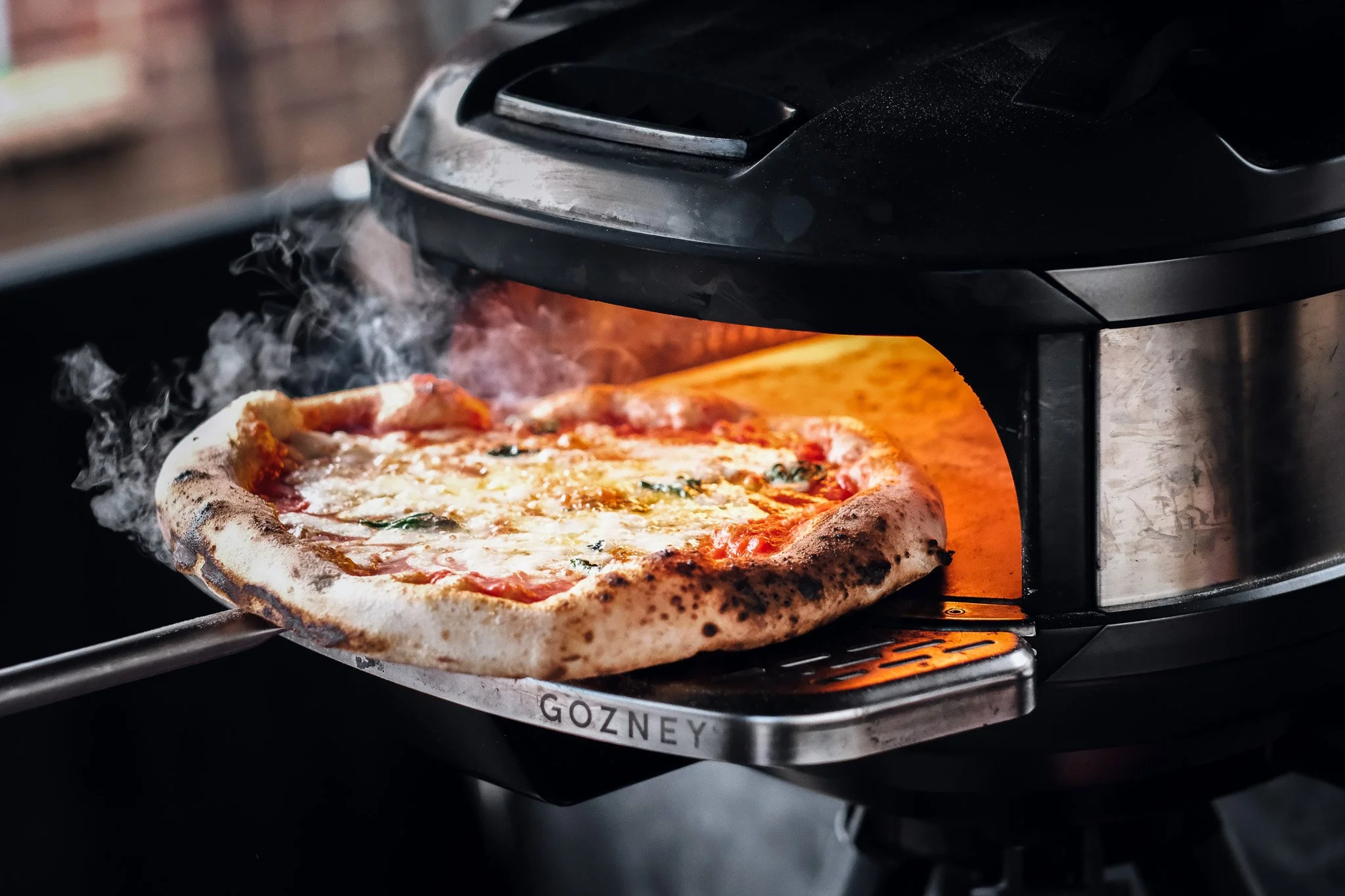 A pizza is being baked in a professional oven with the brand name Gozney on the front. The pizza has cheese, tomato sauce, and herbs.