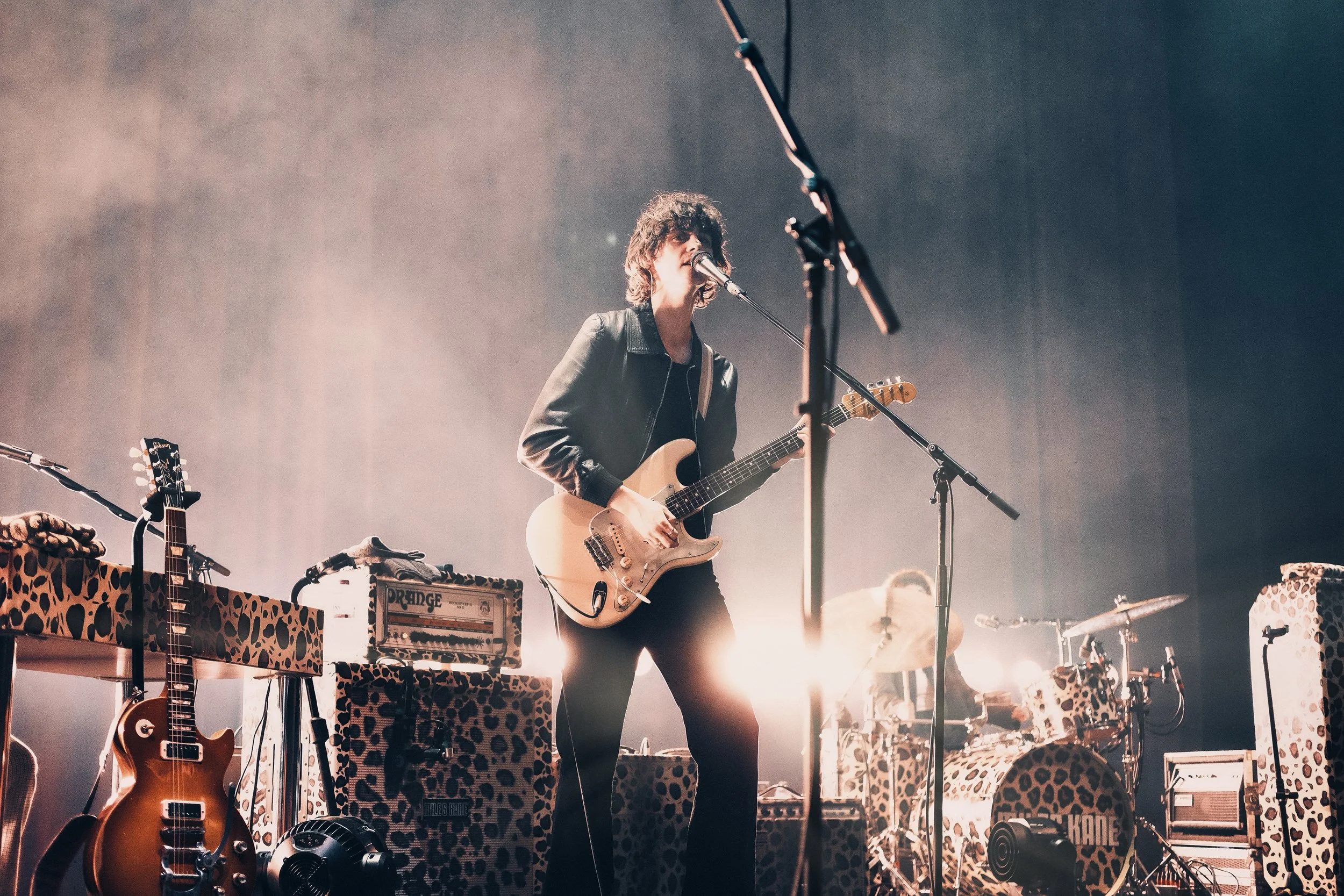 A musician singing into a microphone and playing an electric guitar during a live performance. The stage is decorated with animal print fabric on the equipment and instruments.
