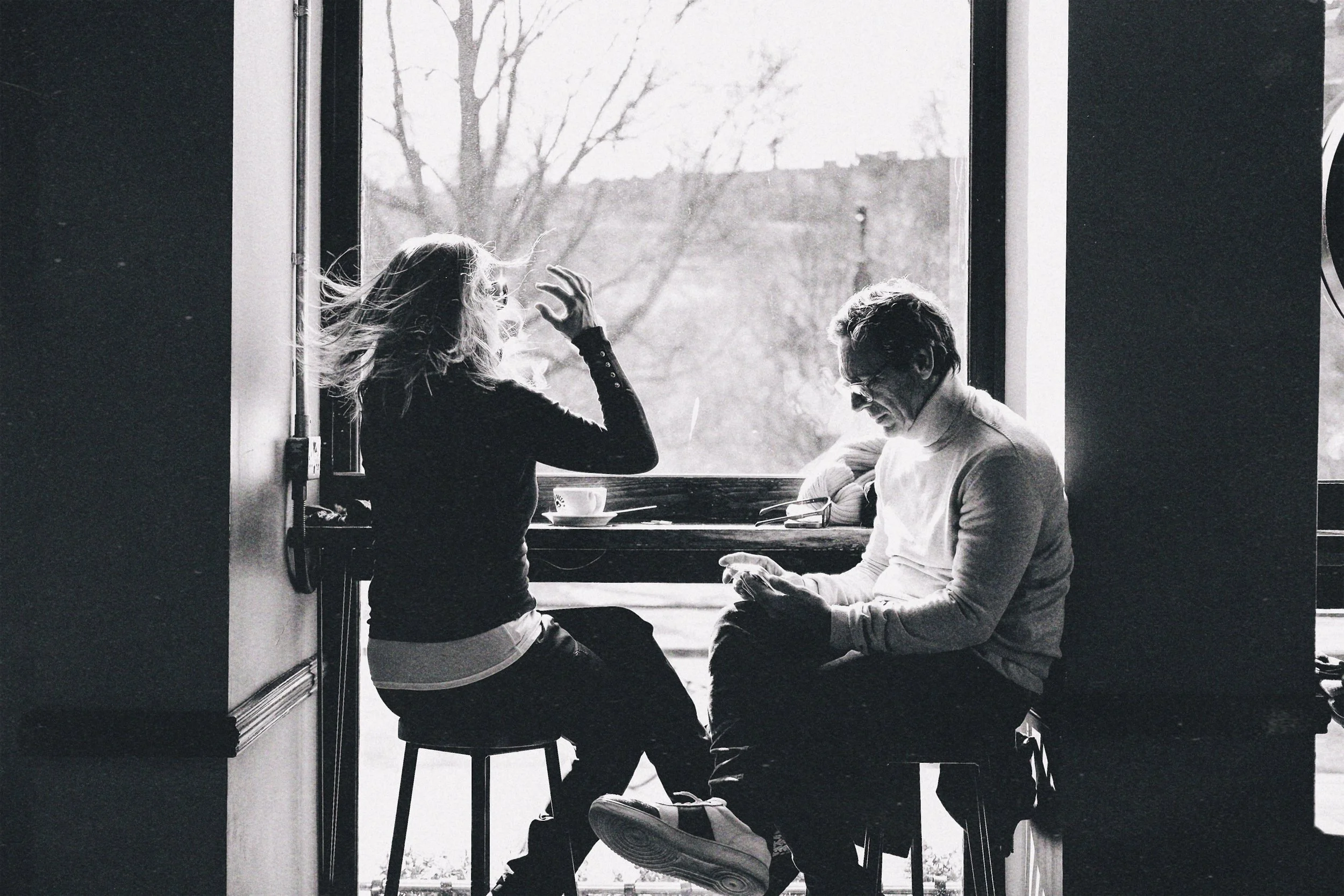 A black and white photo of two people sitting at a window. They are facing each other, with one person gesturing with their hand and the other looking down at a phone. There is a cup on the table and a pair of glasses, with outdoor scenery visible th