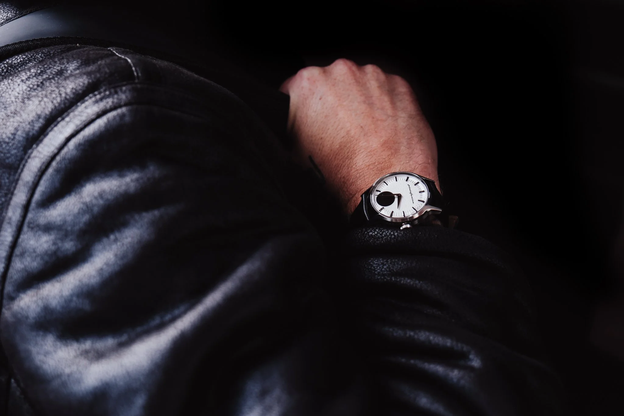 A person's wrist showing a modern wristwatch with a white face and black strap, partially covered by a leather jacket sleeve.