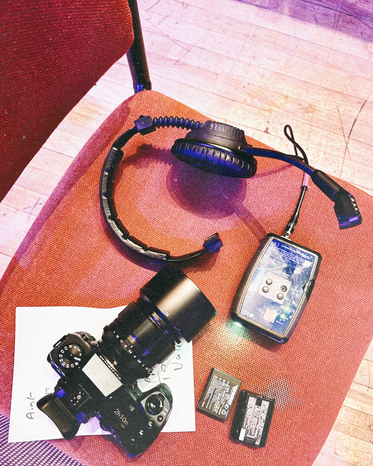 A photographer's live event kit laid out on a venue seat—camera with lens attached, headphones, a wireless monitor unit, and spare batteries, captured under purple stage lighting