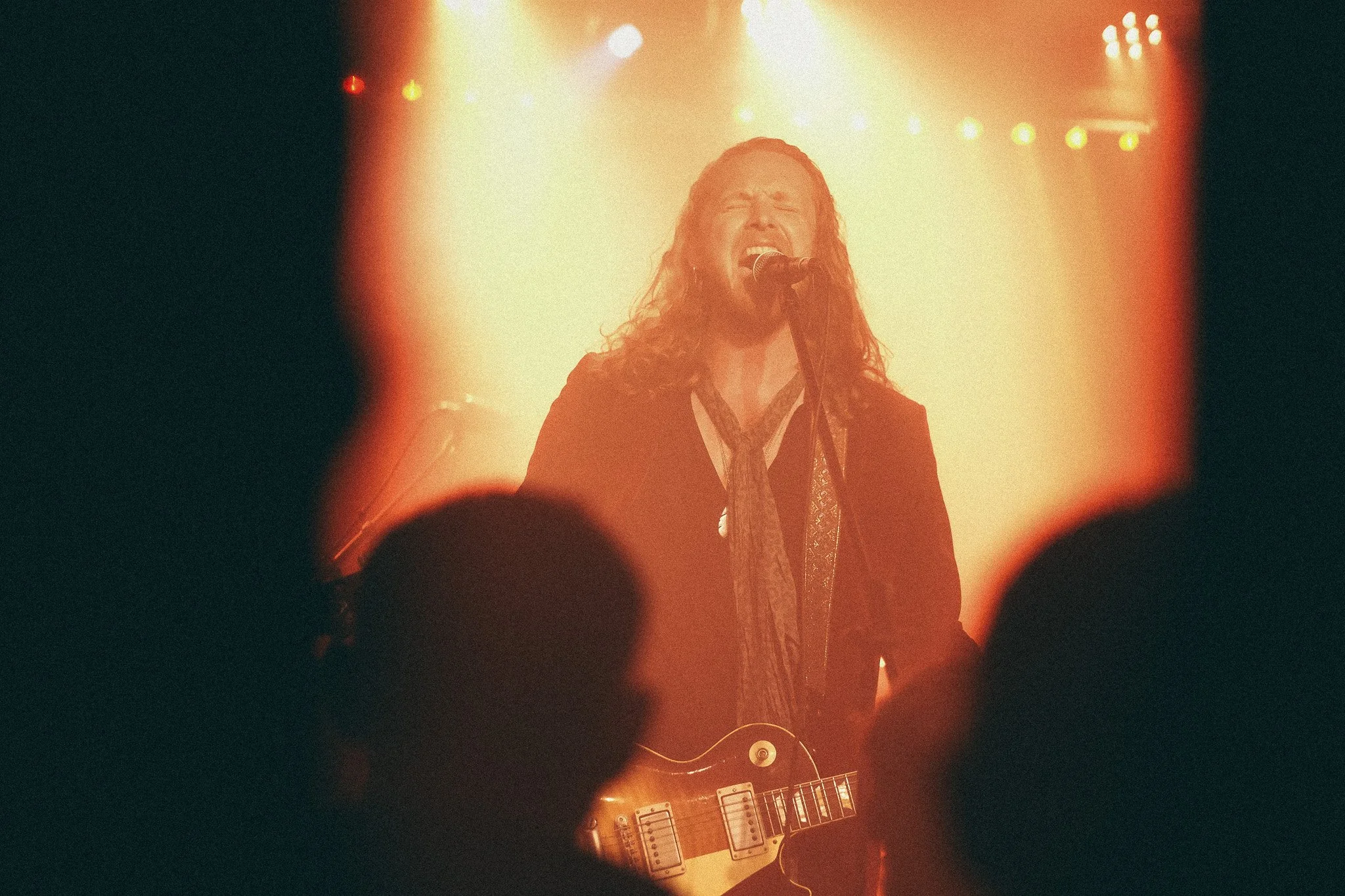 A singer with long hair passionately performing on stage with closed eyes and mouth open, singing into a microphone, illuminated by warm stage lights, with an electric guitar in front of them, viewed through audience members' silhouettes.
