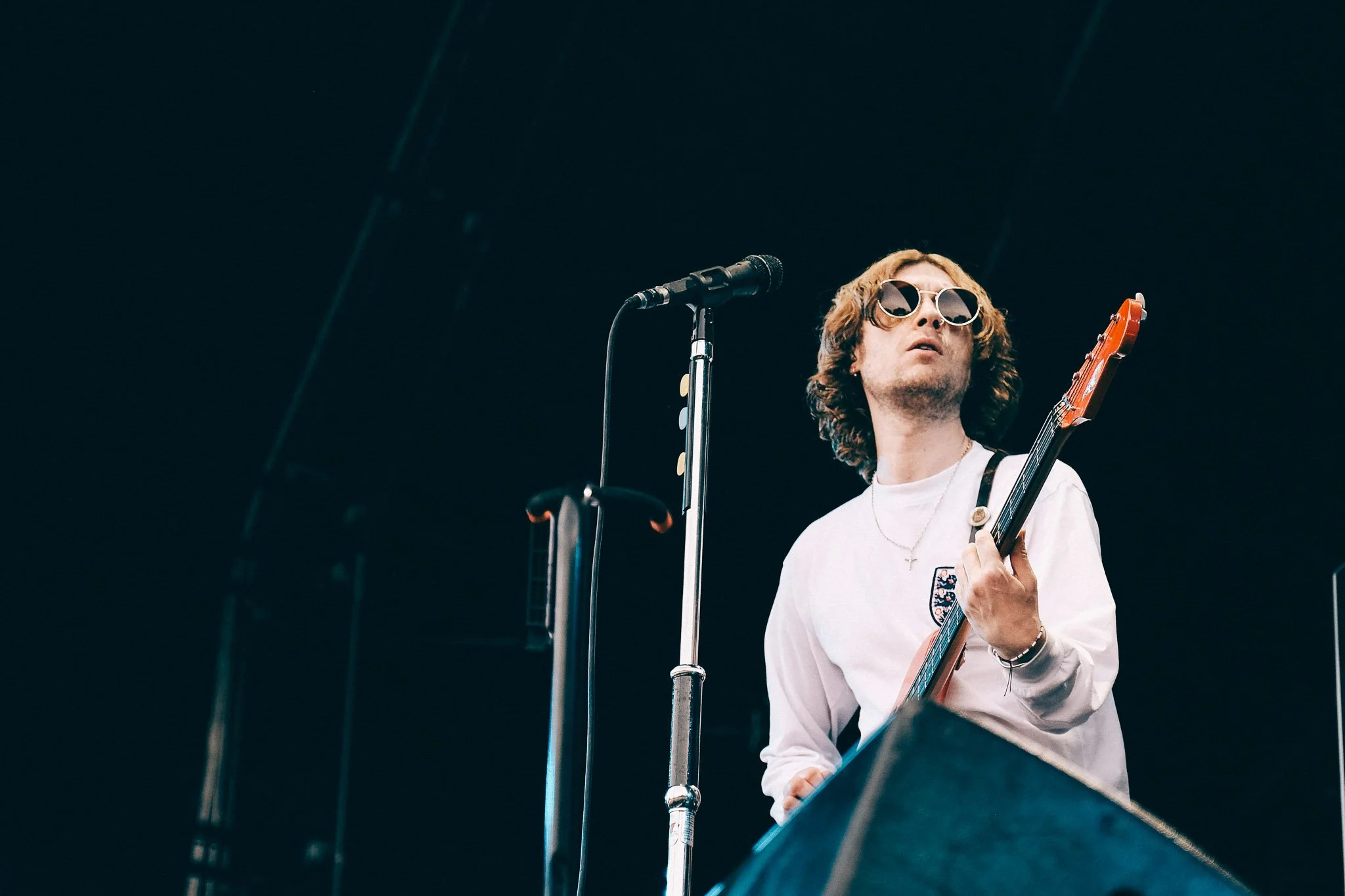 A man wearing sunglasses and a white shirt playing an electric guitar during a performance, with a microphone stand nearby and a dark background.