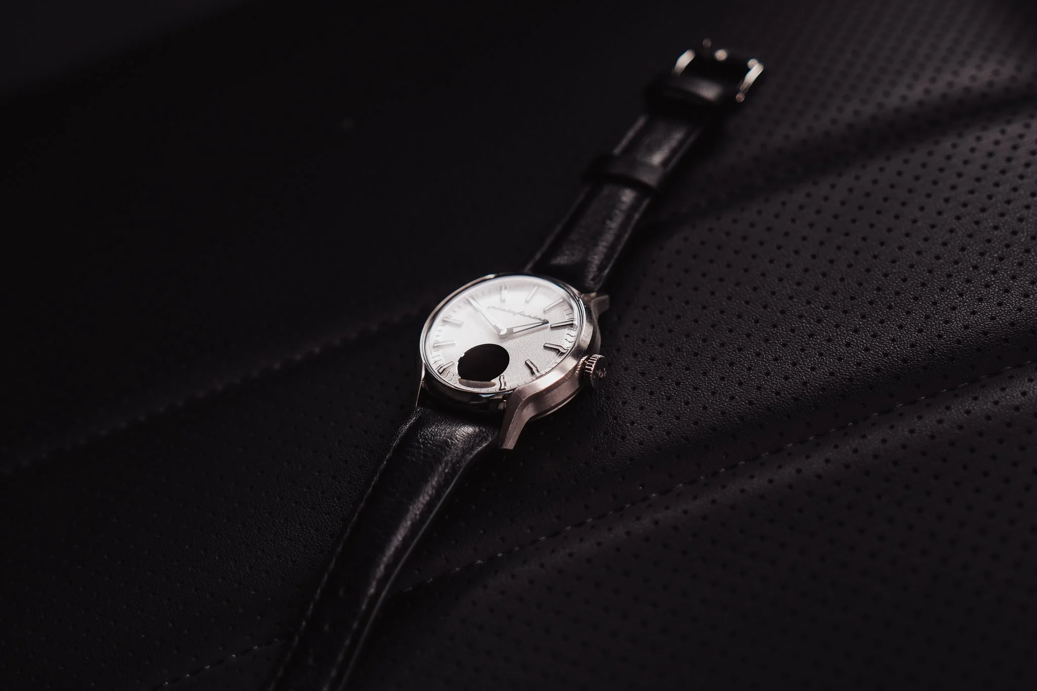 A silver wristwatch with a white face and black leather strap on a black textured surface.