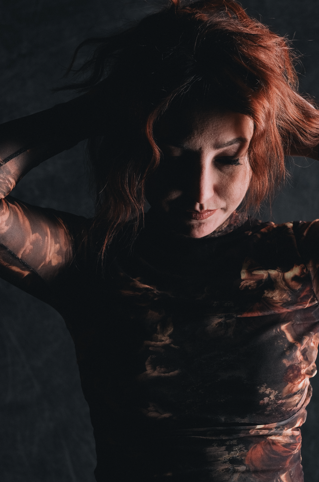 A woman with red hair wearing a sheer, patterned top, holding her head with her hands, looking down with her eyes closed against a dark background.