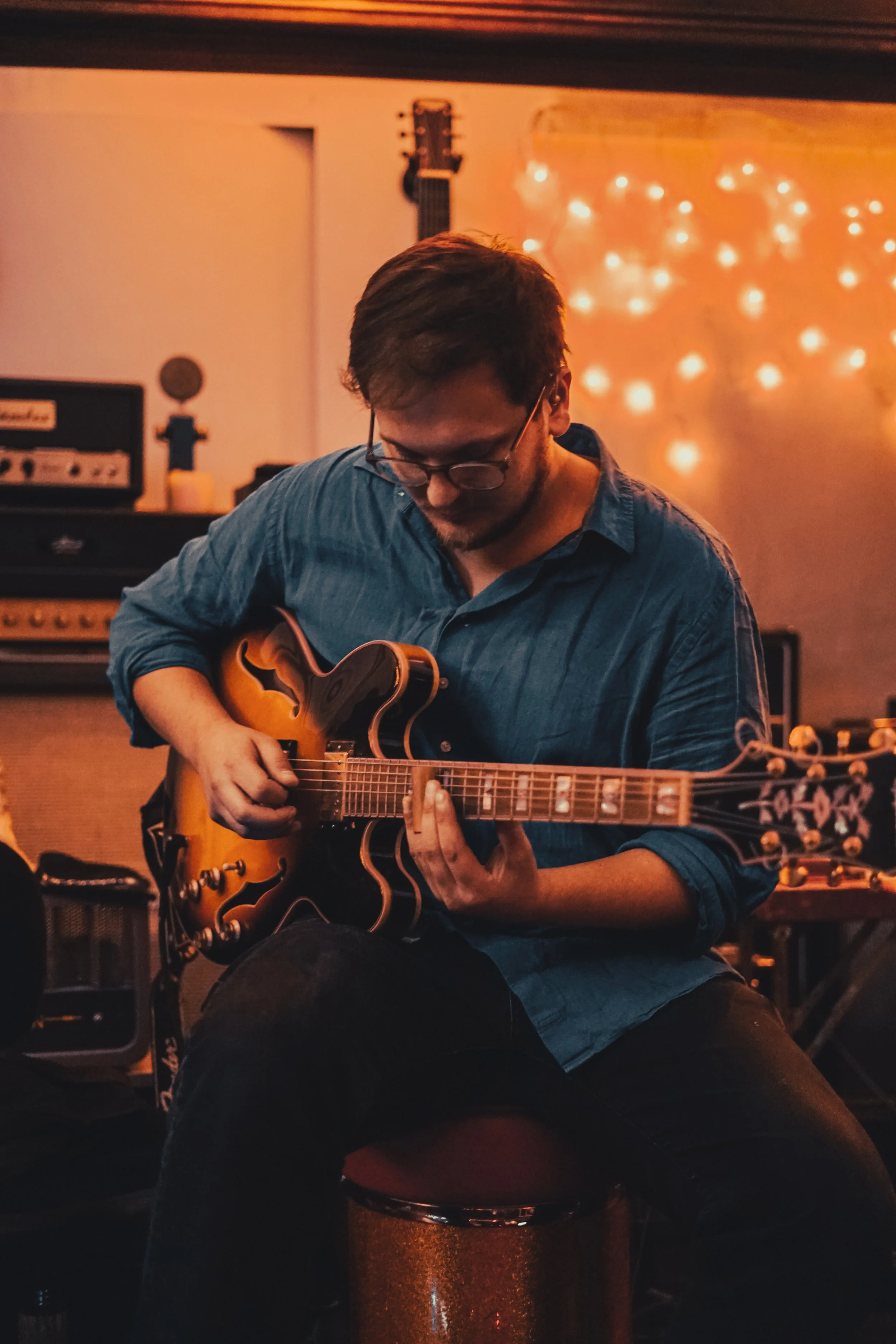 A man with glasses and a blue shirt playing an electric guitar in a cozy, warmly lit room with string lights, music equipment, and an amplifier.