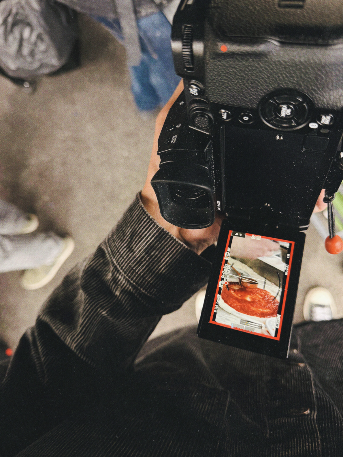 A Fujifilm X-H2 camera held in hand with the flip screen extended, showing live video footage of a food dish being filmed—behind the scenes of a commercial content shoot.