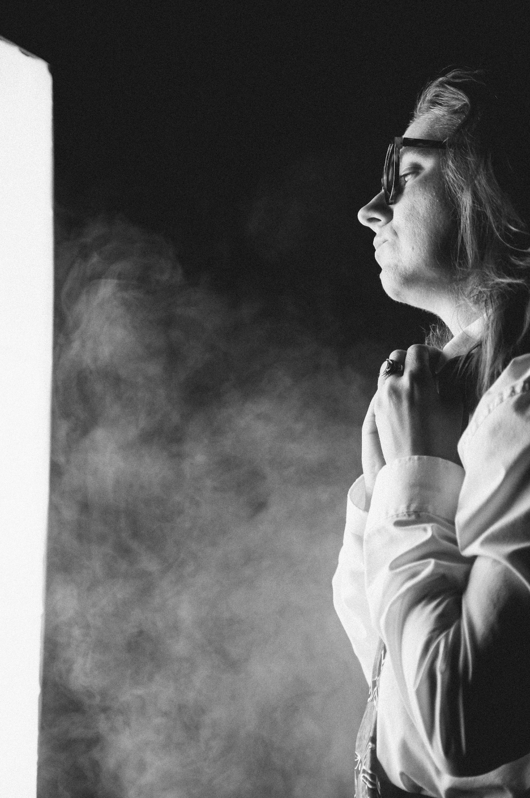 A woman with wavy hair, wearing glasses and a light-colored jacket, looks thoughtfully at a bright screen with clouds of smoke or fog around her. She is holding her hands near her chest.