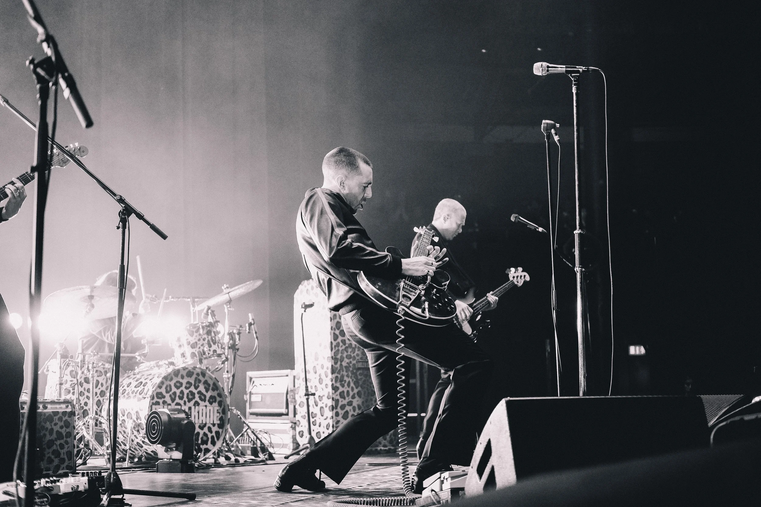 A black and white photo of a band performing on stage, with a lead guitarist in the foreground and a drummer behind. The guitarist is mid-performance, leaning back with his foot forward, playing an electric guitar. Microphones and music equipment are
