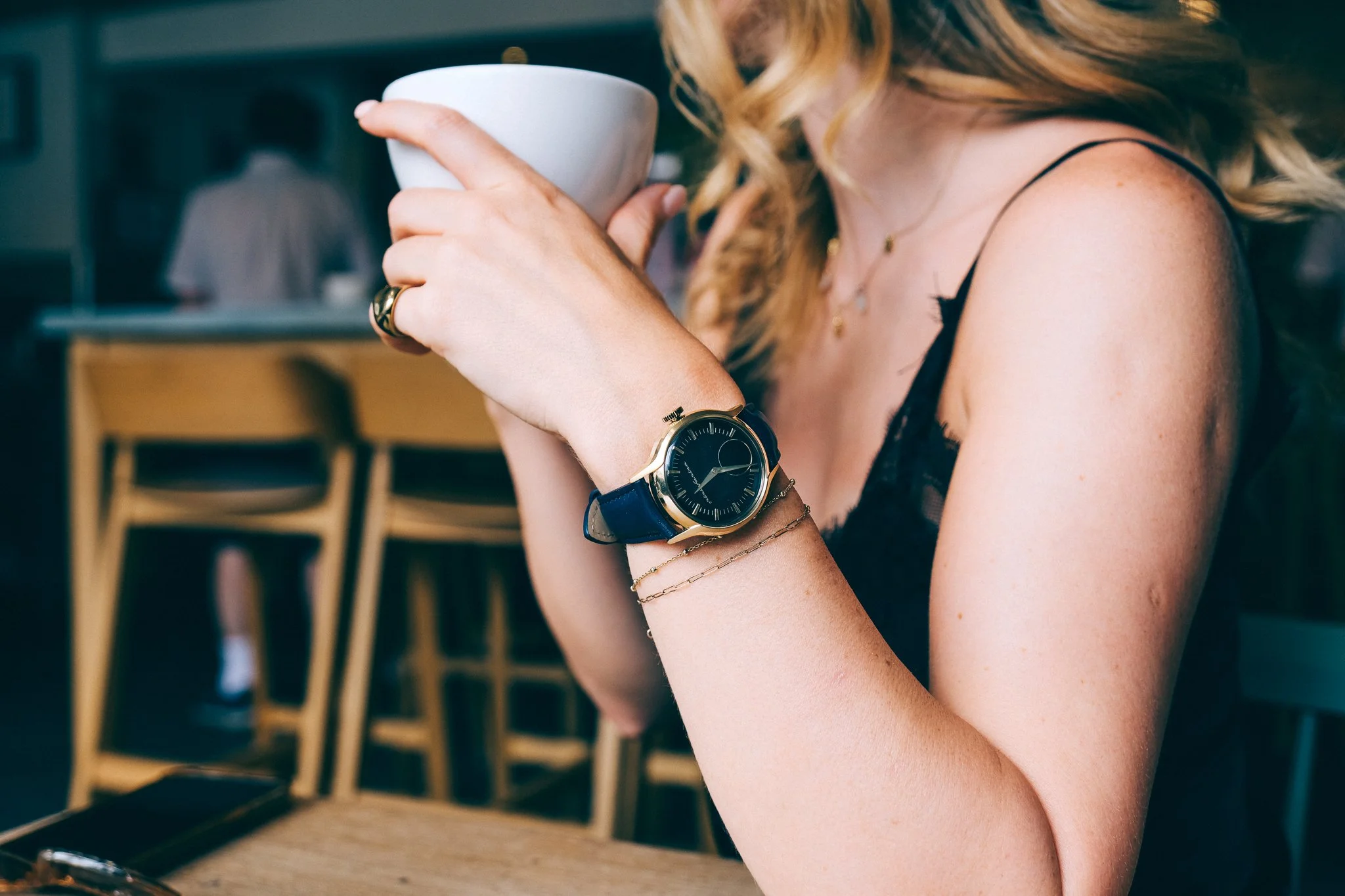 A woman with blonde hair, wearing a black spaghetti strap top, is holding a white mug close to her face. She is wearing a wristwatch, rings, and bracelets, and has layered necklaces, with a blurred background showing a coffee shop or cafe setting.