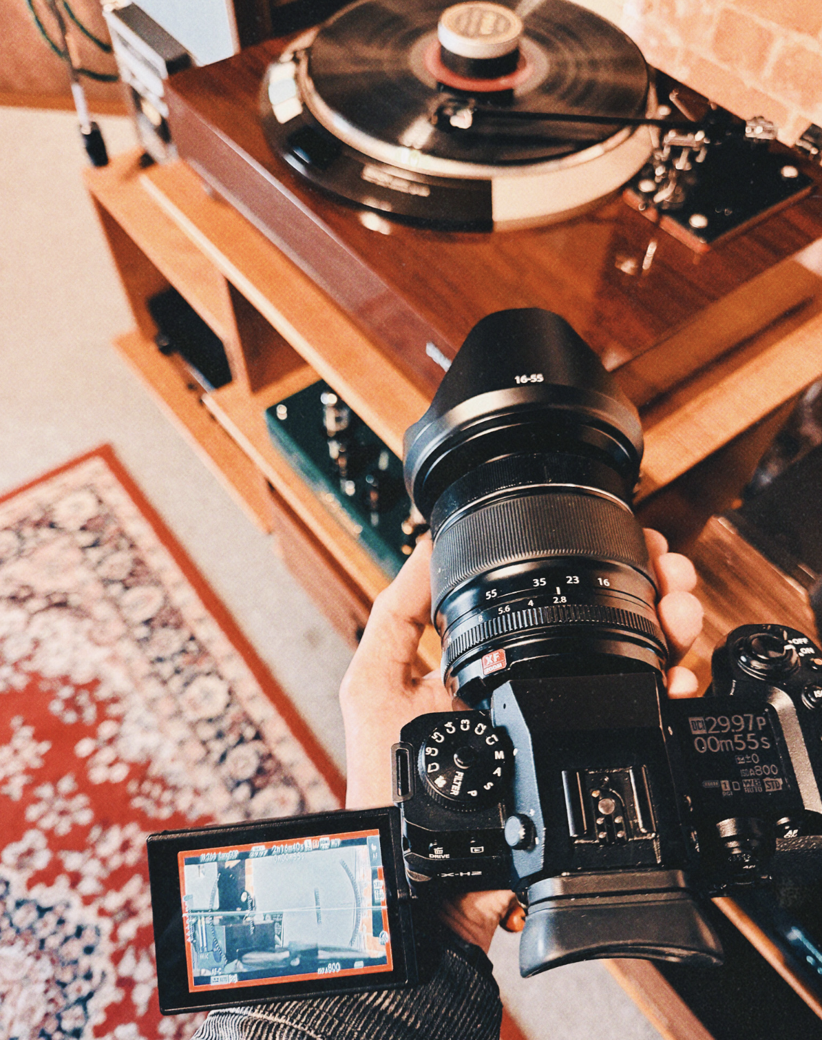 A Fujifilm X-H2 camera held in video recording mode, flip screen extended, framing a vintage vinyl record player—illustrating the hybrid photo and video capabilities of modern cameras.