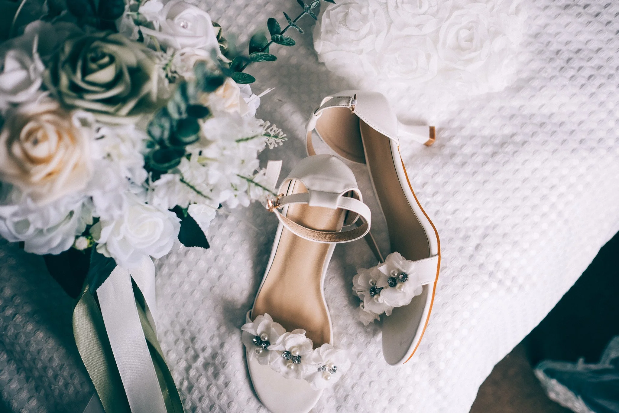 A pair of white high-heeled shoes with decorative pearls and flowers, placed on a textured white surface, next to a bouquet of white and pastel roses and greenery.