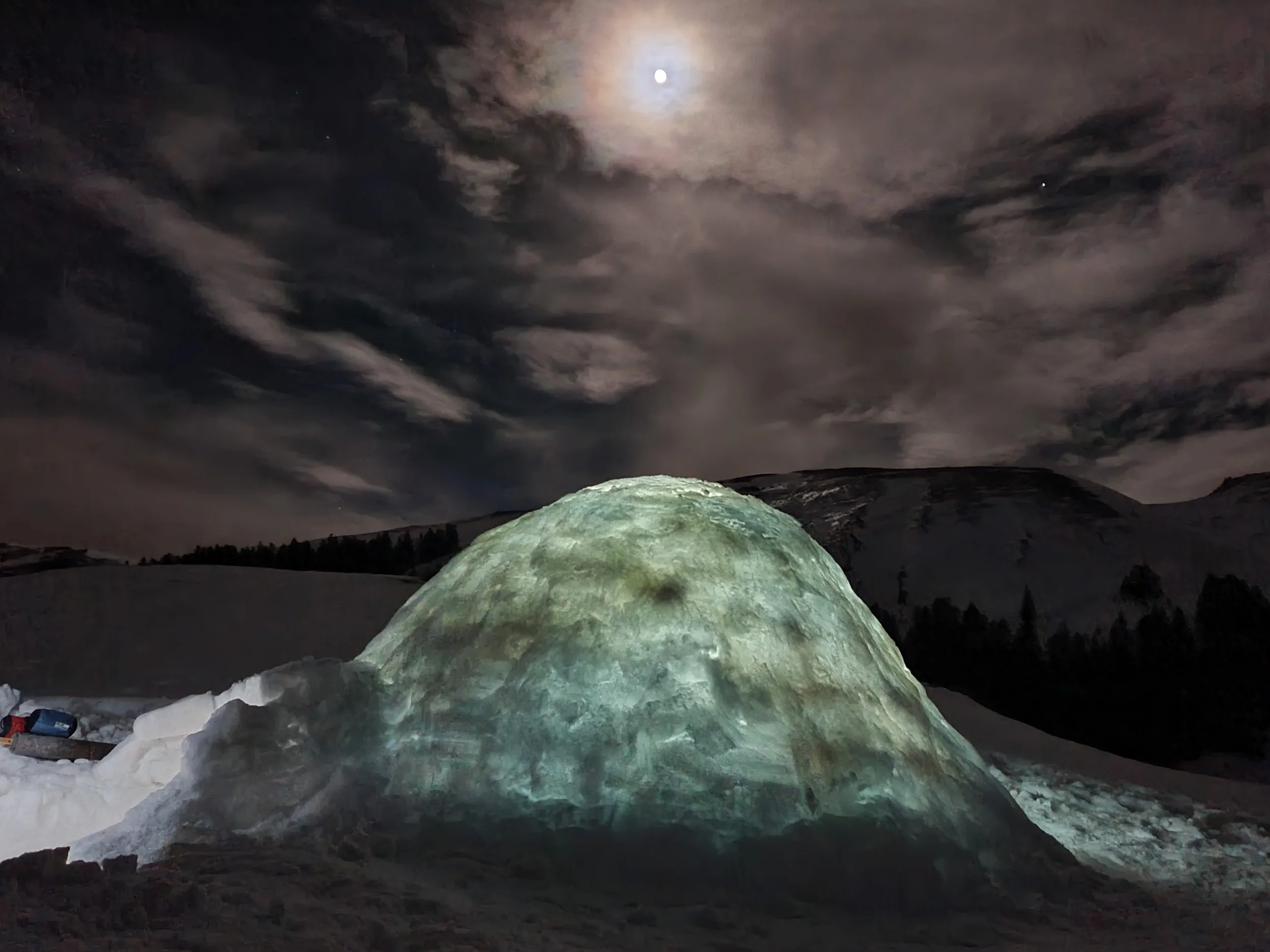 Morceau de glace illuminé sous la lune dans un paysage montagnard nocturne avec des nuages dans le ciel.
