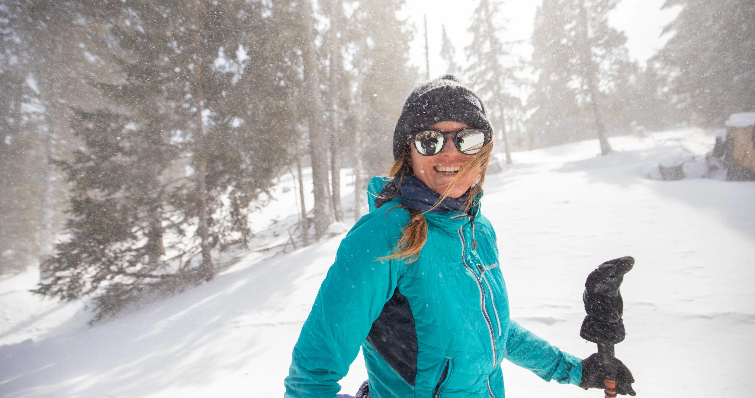 Femme souriante en tenue de ski, portant des lunettes de soleil, une veste bleue, un bonnet noir et des gants noirs, dans un paysage enneigé avec des arbres et de la neige tombante.