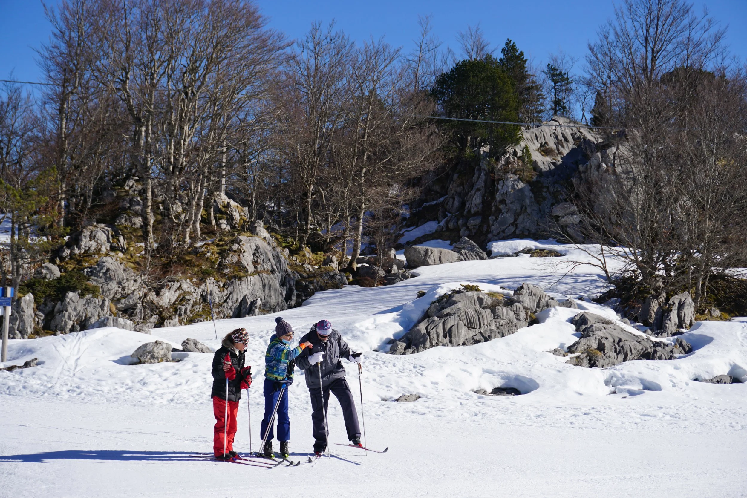 Trois personnes en vêtements de ski discutent sur une surface enneigée avec un paysage rocheux et des arbres dénudés en arrière-plan, sous un ciel bleu.
