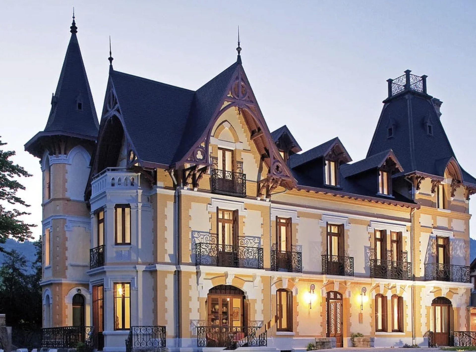 Une grande maison de style victorien avec des tourelles, des balcons en fer forgé et des fenêtres illuminées, prise au crépuscule.