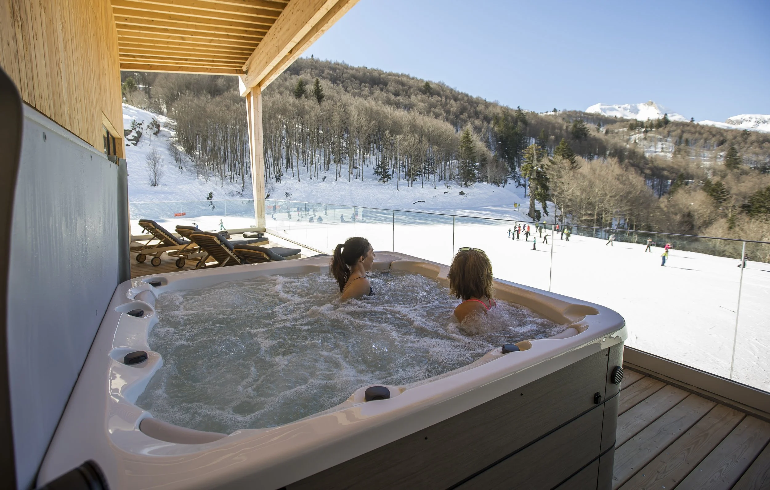 Deux femmes relaxant dans un jacuzzi sur une terrasse avec vue sur une station de ski enneigée, avec des skieurs en arrière-plan.