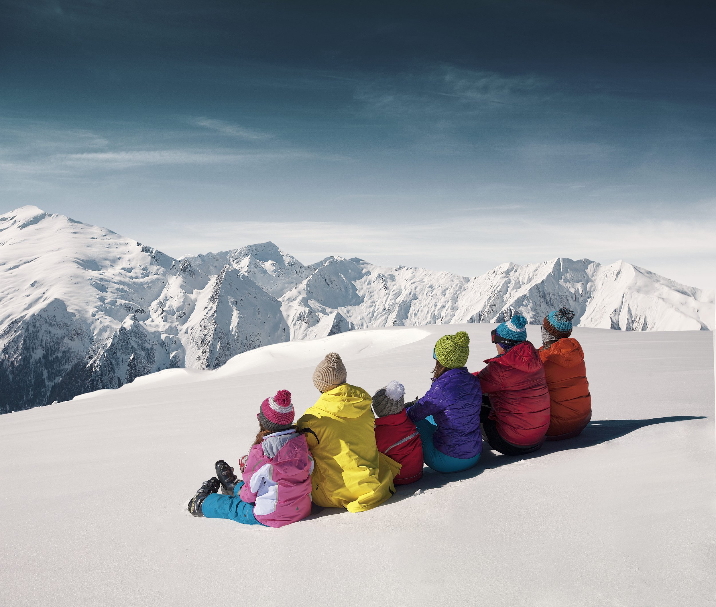 Groupe d'enfants en vêtements d'hiver assis dans la neige, regardant une chaîne de montagnes enneigées sous un ciel bleu.