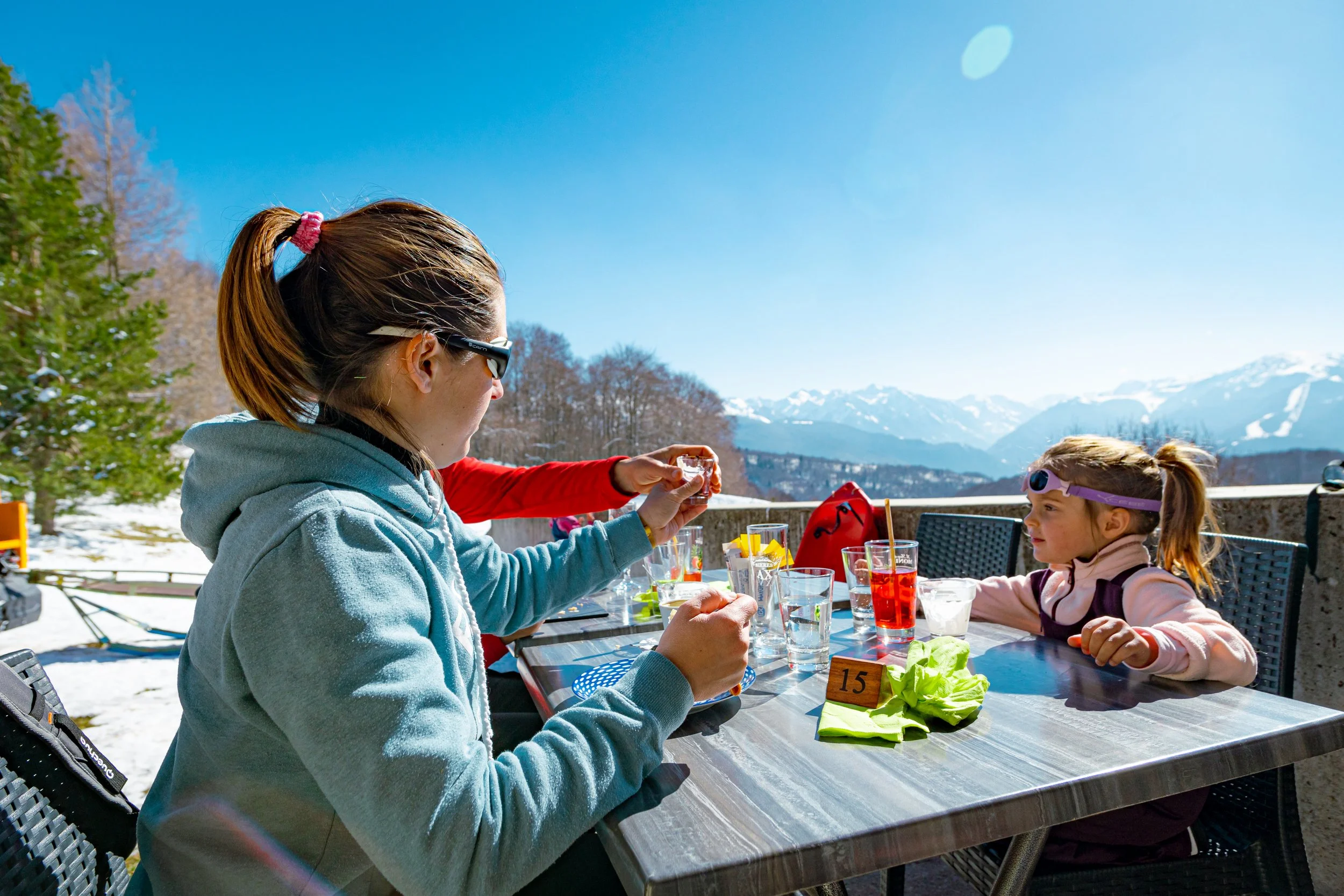 Deux femmes et une fille jeune s'assoient à une table en plein air dans un paysage montagnard enneigé, buvant des boissons et profitant du beau temps. La scène se déroule sous un ciel bleu clair.