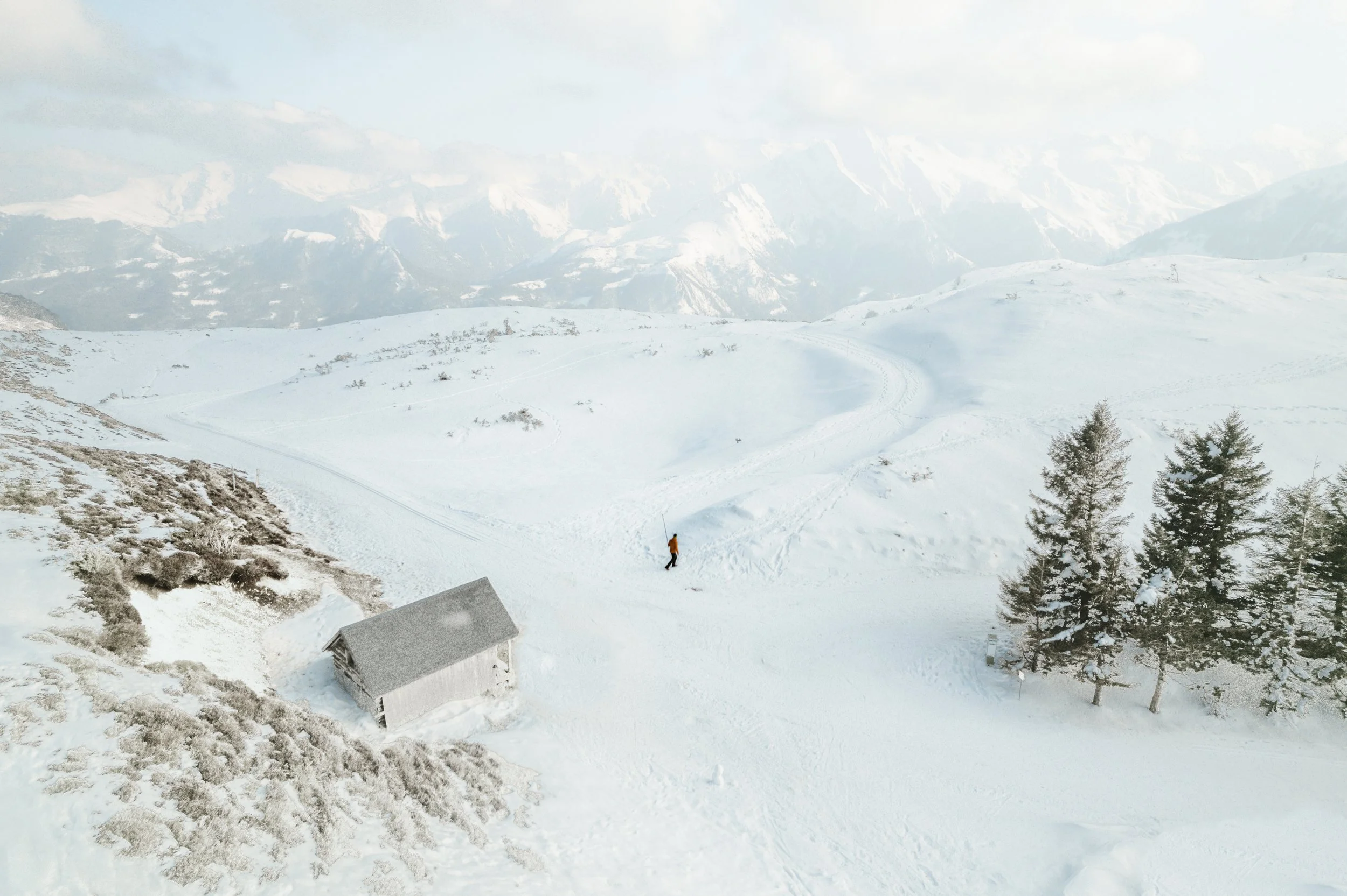 Paysage de montagne enneigée avec un petit chalet et un skieur dans la neige, arbres et montagnes en arrière-plan.