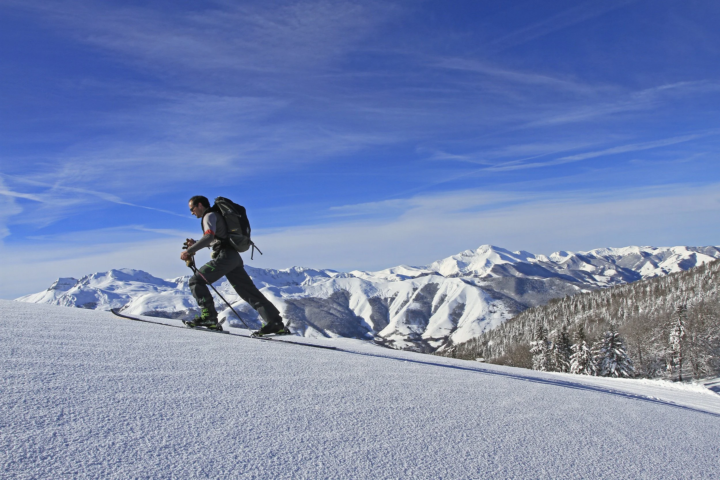 Un skieur soliste avec un sac à dos qui descend une pente enneigée avec une montagne de sommets enneigés en arrière-plan sous un ciel bleu.