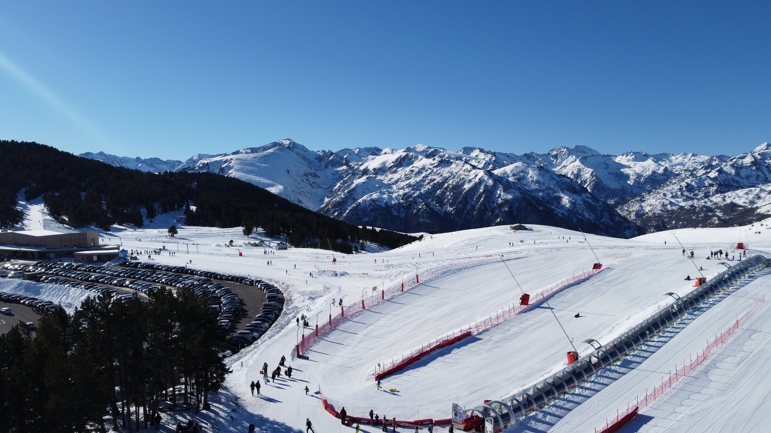 Vue d'une station de ski avec des skieurs, un parking rempli de voitures, des montagnes enneigées en arrière-plan, et une piste de ski couverte de neige.