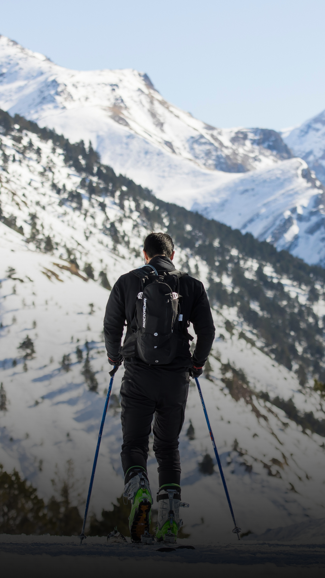 Un homme en tenue de ski avec un sac à dos et des bâtons de ski ascendante dans un paysage de montagnes enneigées.