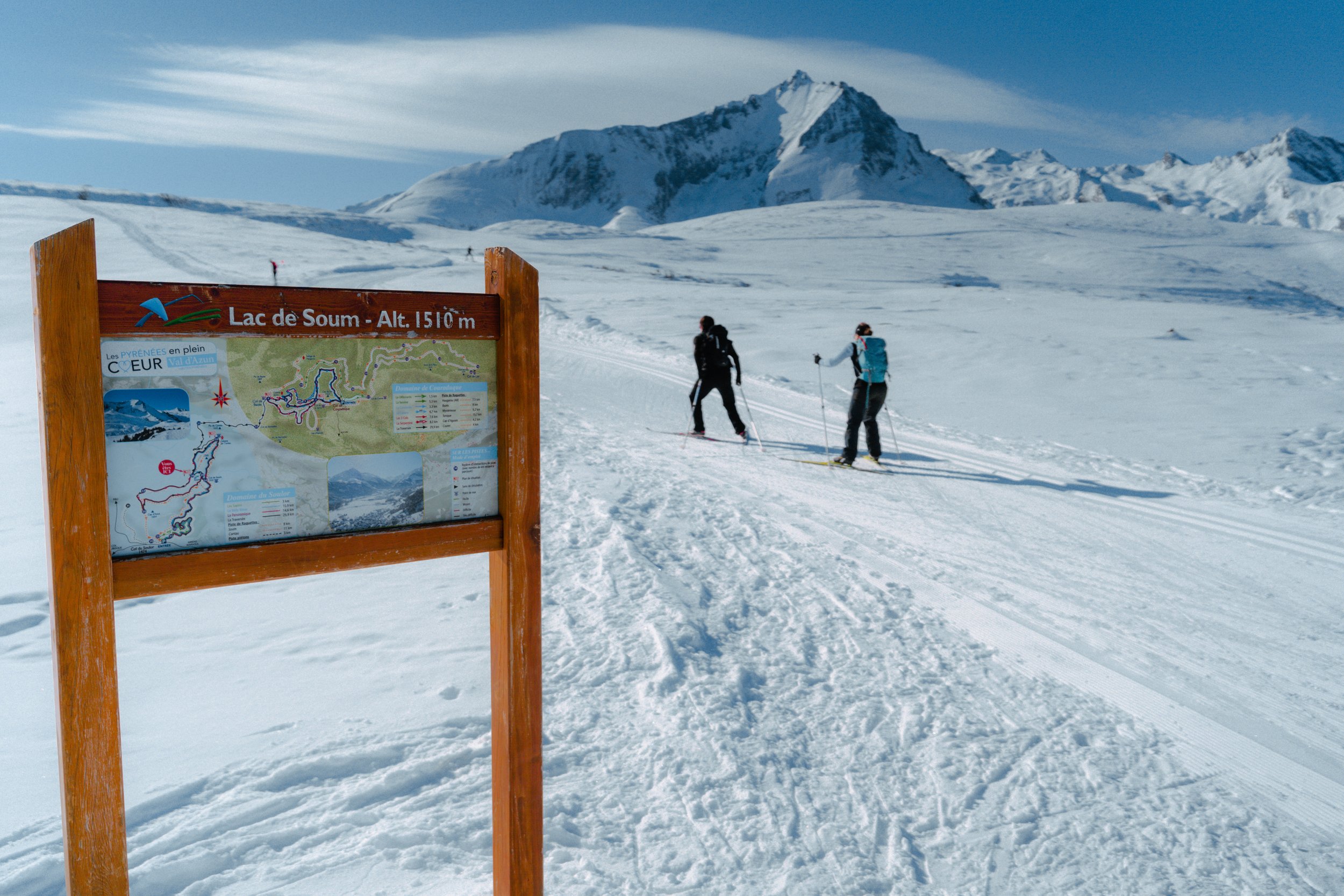 Pistes de ski dans un paysage enneigé dans les Pyrénées, avec un tableau d'information en premier plan, monts enneigés en arrière-plan, deux skieurs en milieu de parcours.
