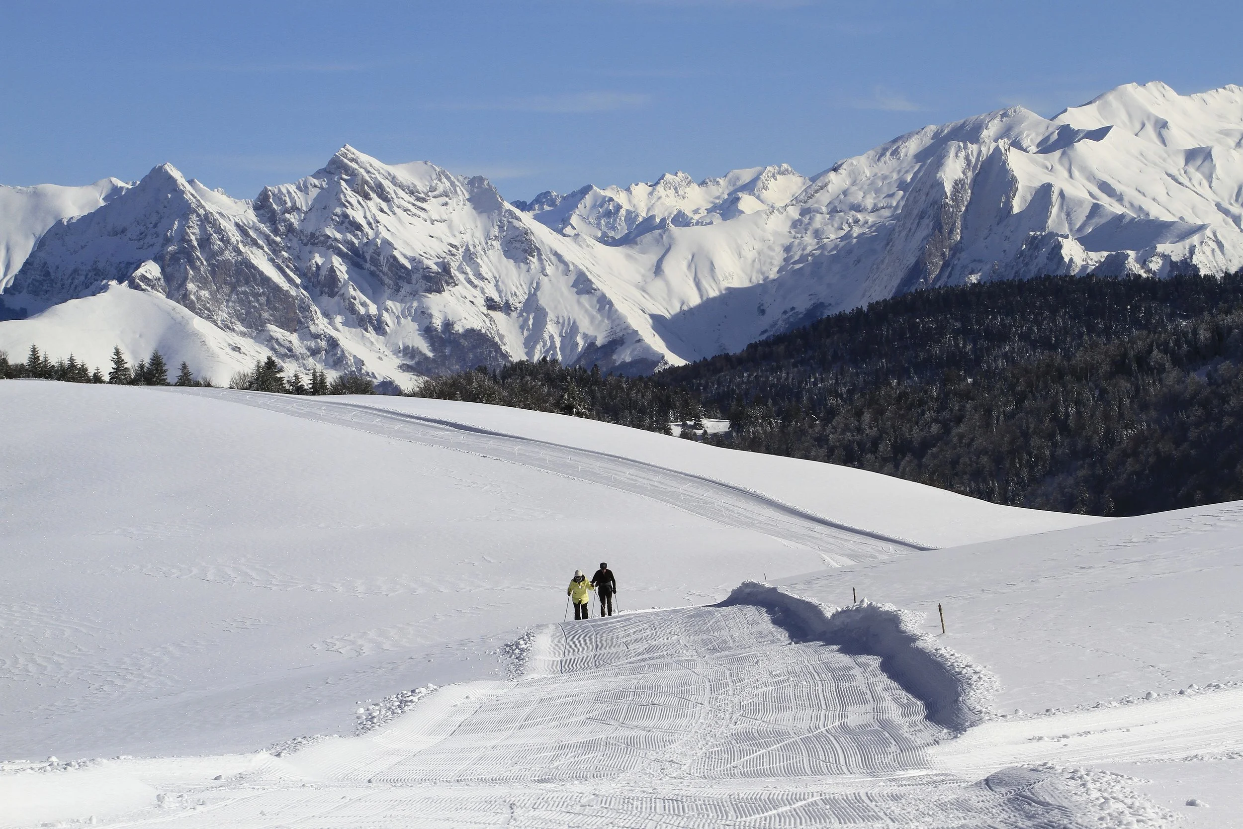 Deux personnes faisant du ski de fond dans un paysage enneigé avec des montagnes imposantes en arrière-plan, ciel bleu clair.