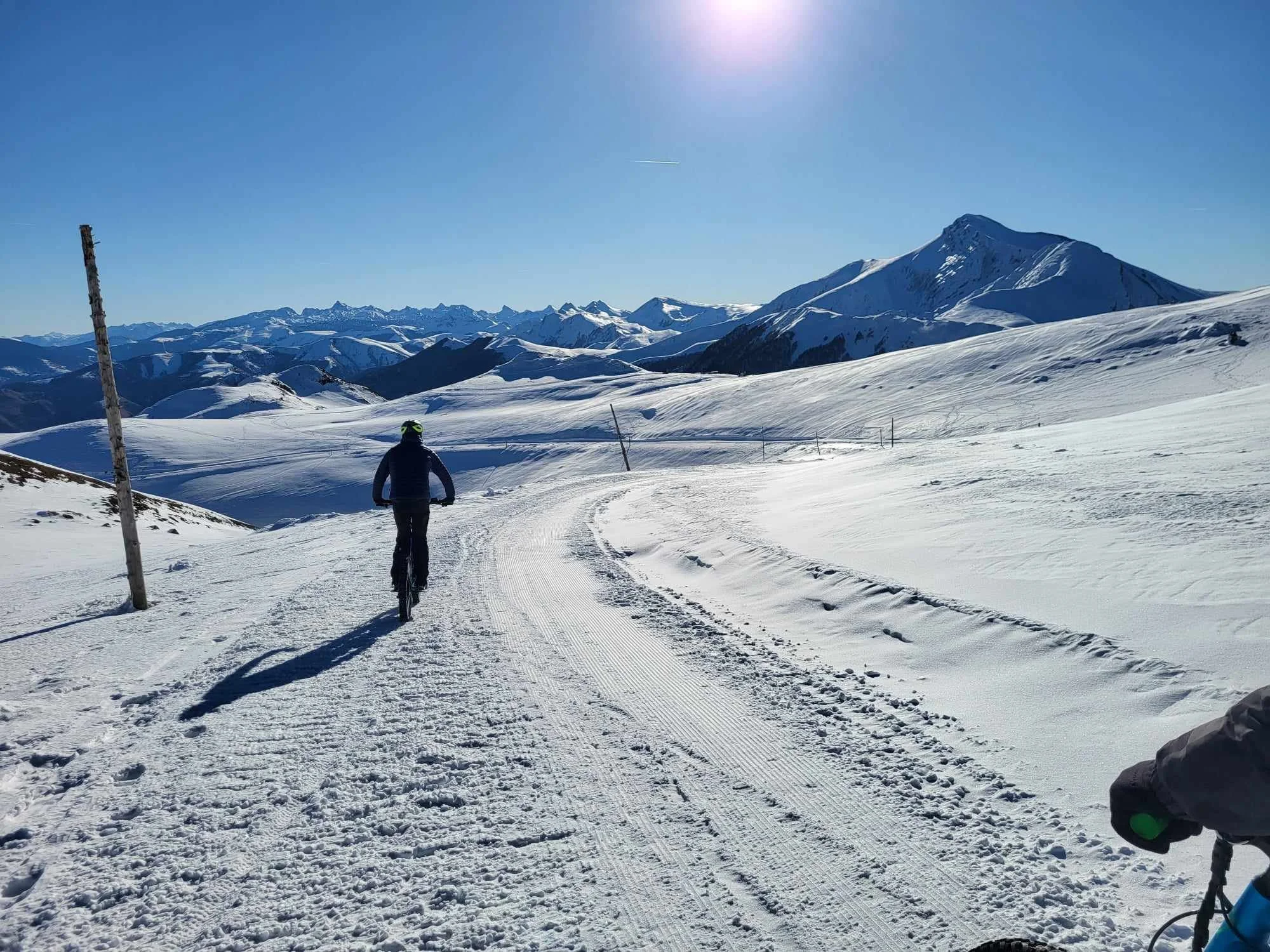 Personne faisant du vélo en montagne enneigée sous un ciel clair, avec des montagnes et un sommet enneigé à l'arrière-plan.