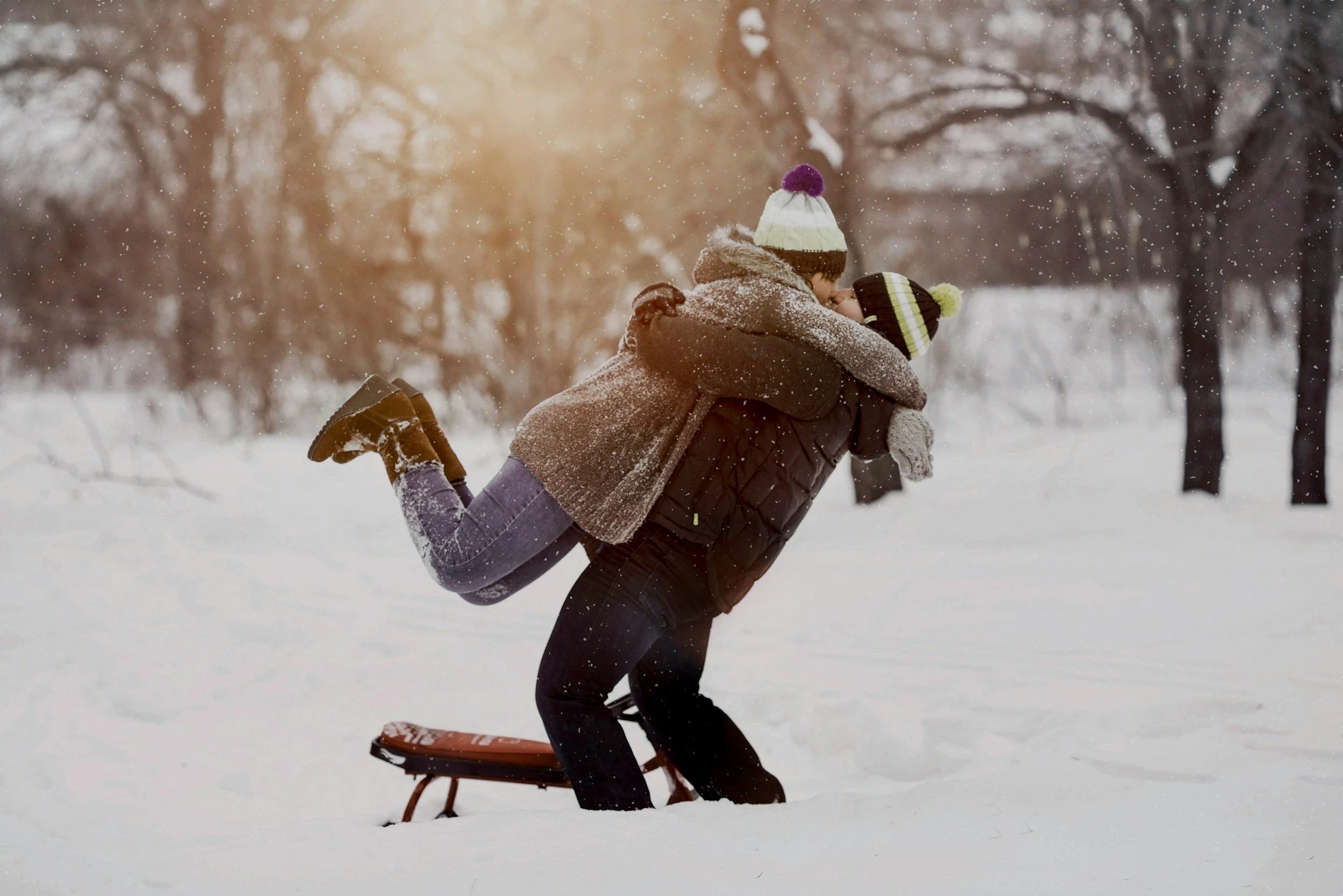 Deux personnes jouent dans la neige, dont une personne donne un coup de pied à une autre, dans un parc enneigé avec des arbres en arrière-plan.