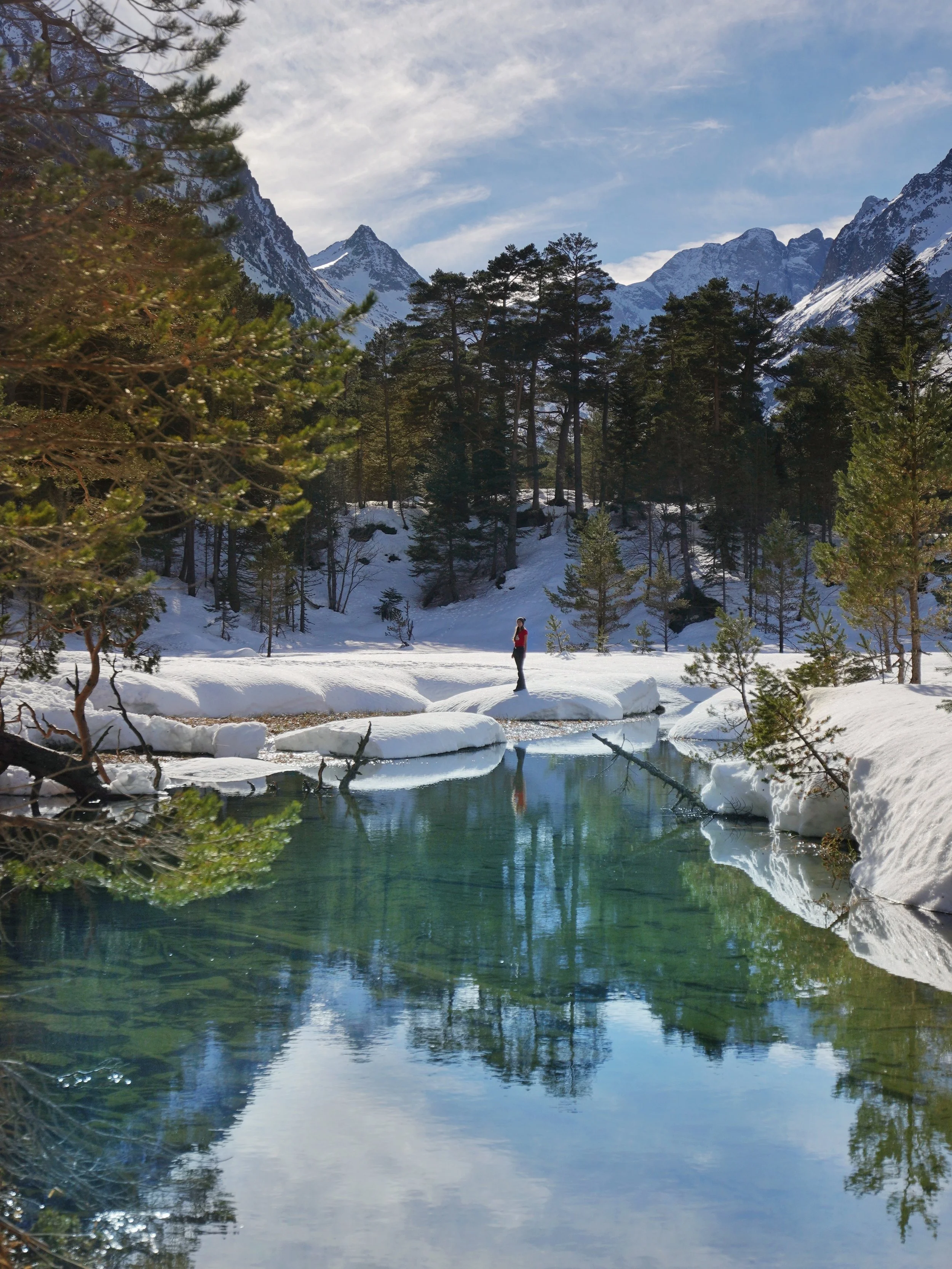 Une personne debout sur un rocher au bord d'un lac glacé entouré de forêts et de montagnes enneigées, sous un ciel partiellement nuageux.