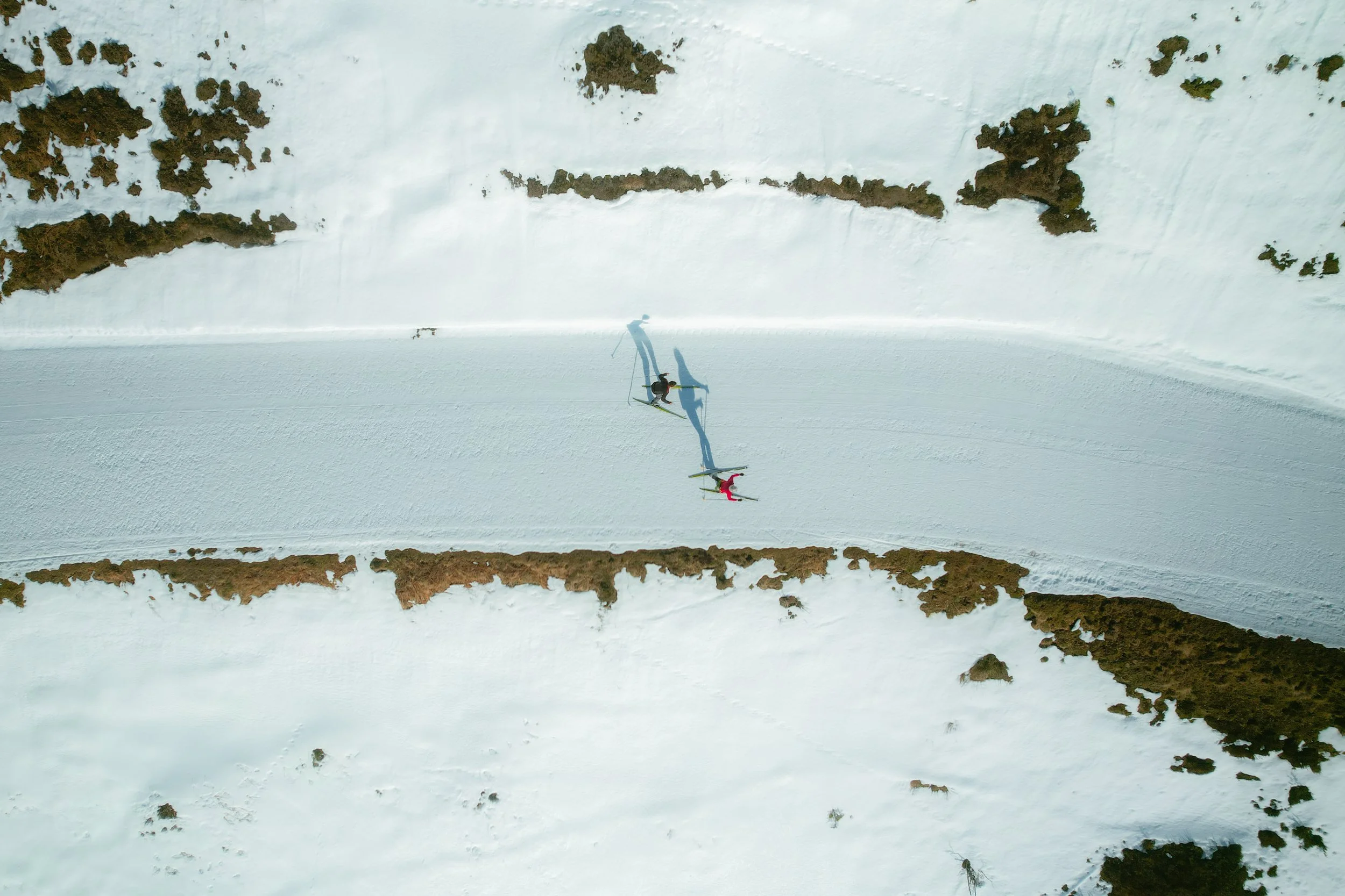 Deux skieurs descendent une piste enneigée dans un paysage de montagne avec de la neige et quelques arbustes ou rochers.