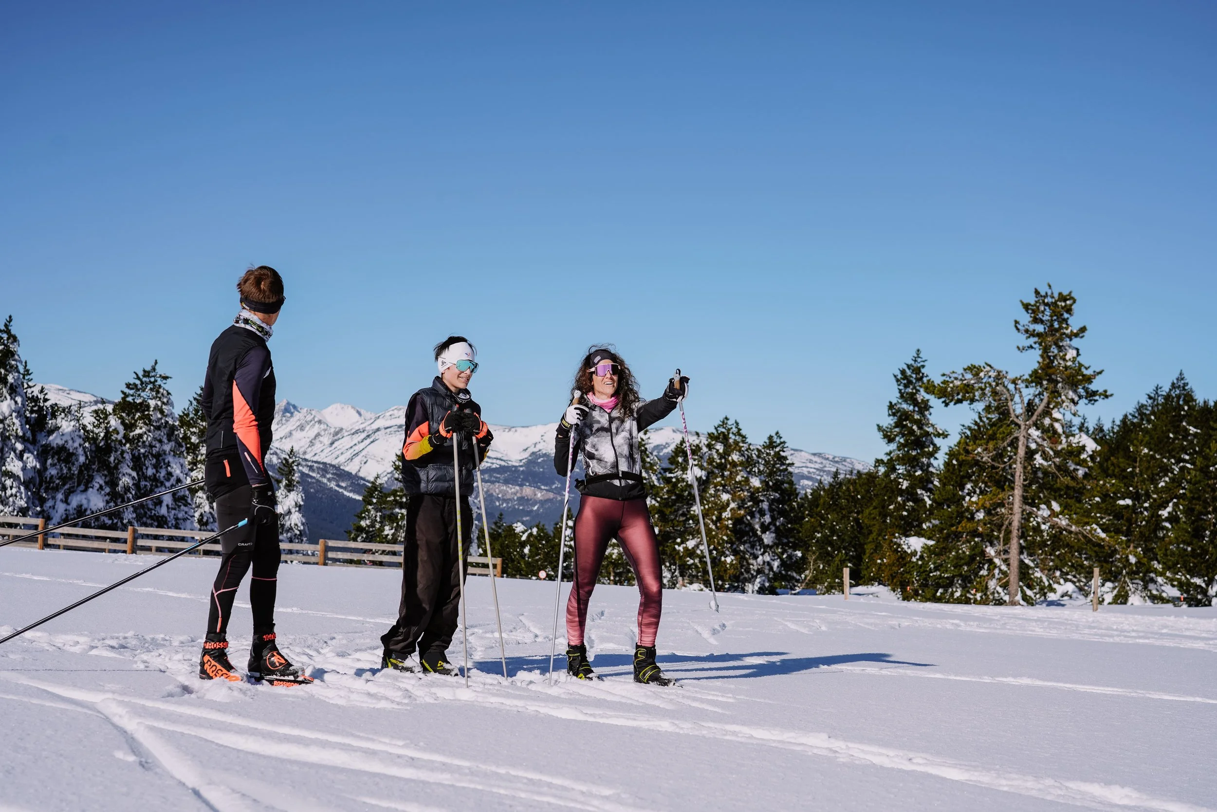 Trois personnes en ski dans un paysage enneigé avec des montagnes et des arbres, ciel bleu.